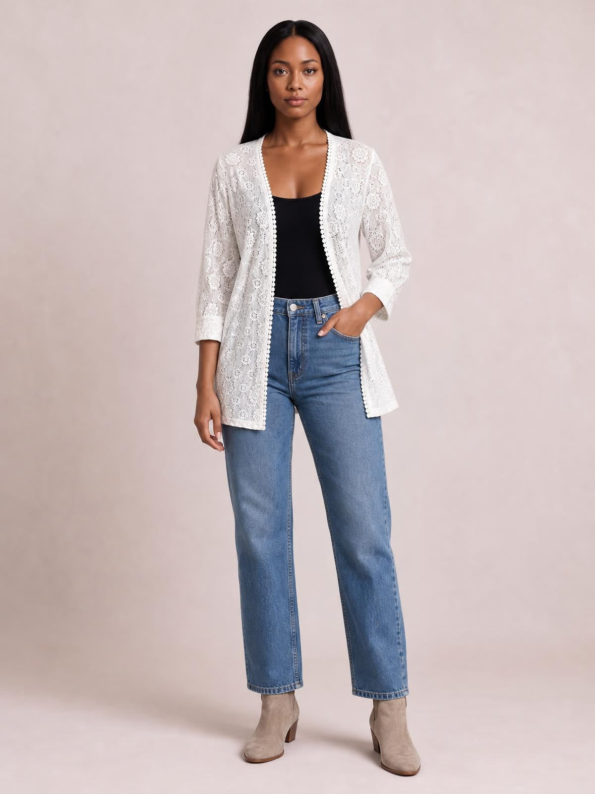 A model stands in a white lace open-front cover-up with long sleeves, scalloped edging, and a relaxed longline silhouette over a black top, one hand in her jeans pocket.