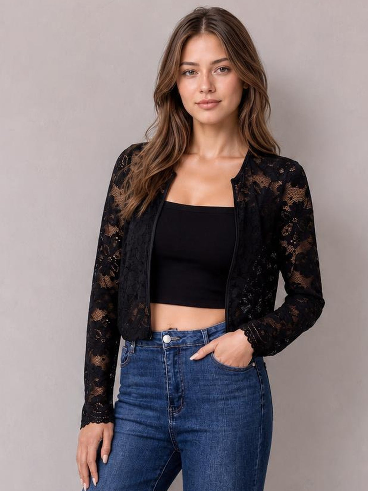 Model wearing a black lace open front long sleeve jacket with a fitted, waist-length silhouette and floral construction, posing with one hand in her jeans pocket.