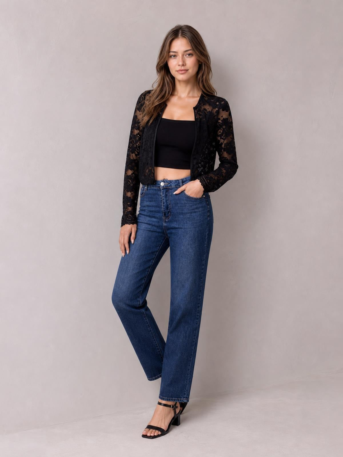 A model poses in a black lace open front long sleeve jacket with a fitted waist-length silhouette, standing with one hand in her jeans pocket.