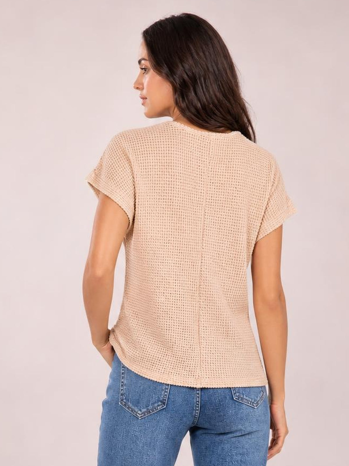 A model shown from behind wears a beige waffle-knit short-sleeve top with a relaxed silhouette, rolled sleeve cuffs, and a gently curved hem.