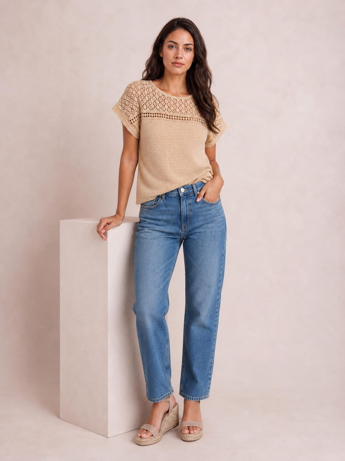 A model wears a beige lace patchwork waffle knit top with short sleeves and a relaxed silhouette, posing with one hand in her jeans pocket.