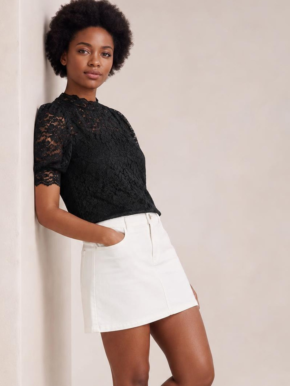 A model leans with one hand in her pocket wearing a black lace top with a regular fit, mock neck, short puff sleeves, and scalloped edges.