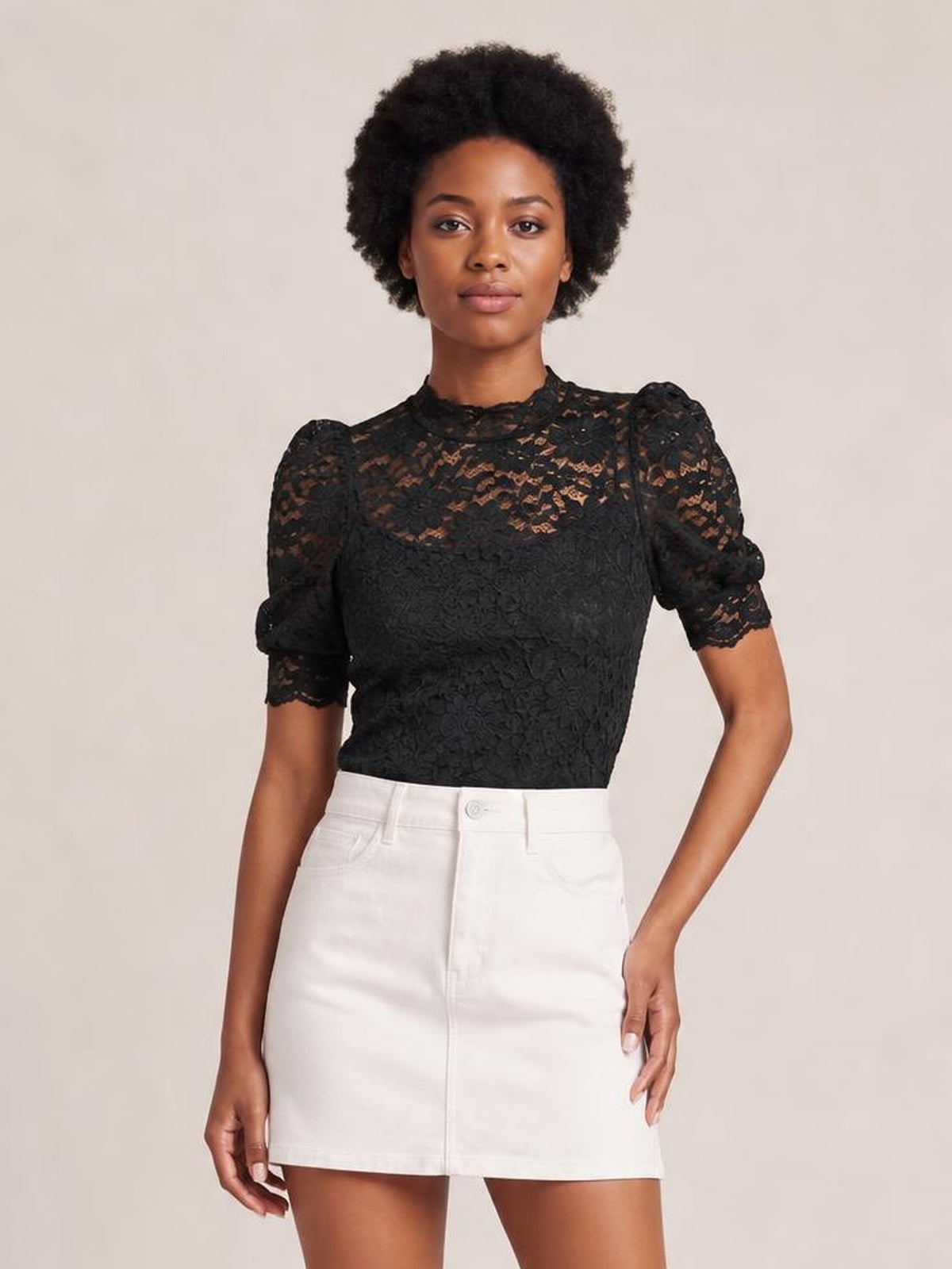 A model stands wearing a black lace top with a mock neck, scalloped trim, short puff sleeves, and a fitted regular silhouette.