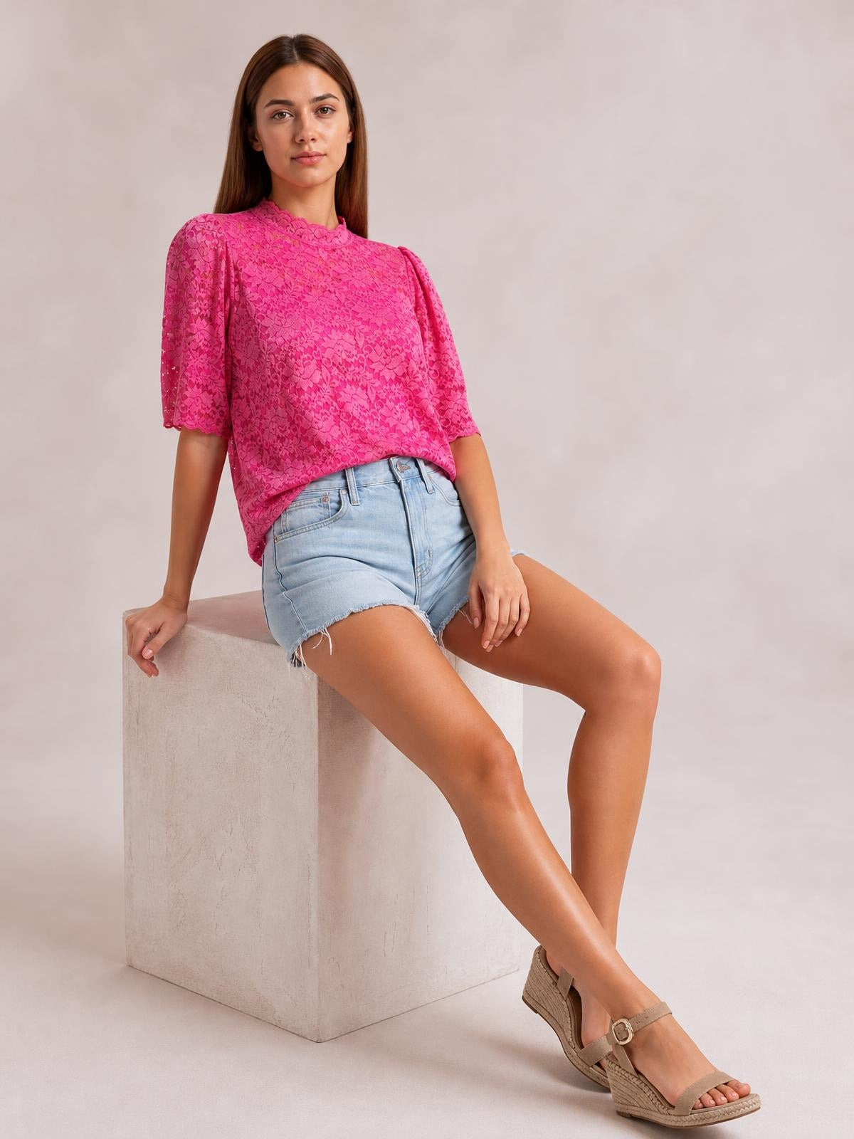 A seated model wears a bright pink lace top with a scalloped mock neck, short puff sleeves, and a relaxed regular-fit silhouette.