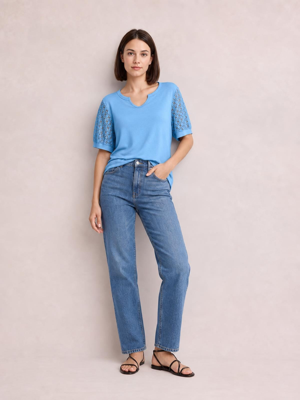 Model wears a blue lace short sleeve T-shirt with semi-sheer openwork sleeves, a relaxed silhouette, and a notched neckline while standing with one hand in her pocket.