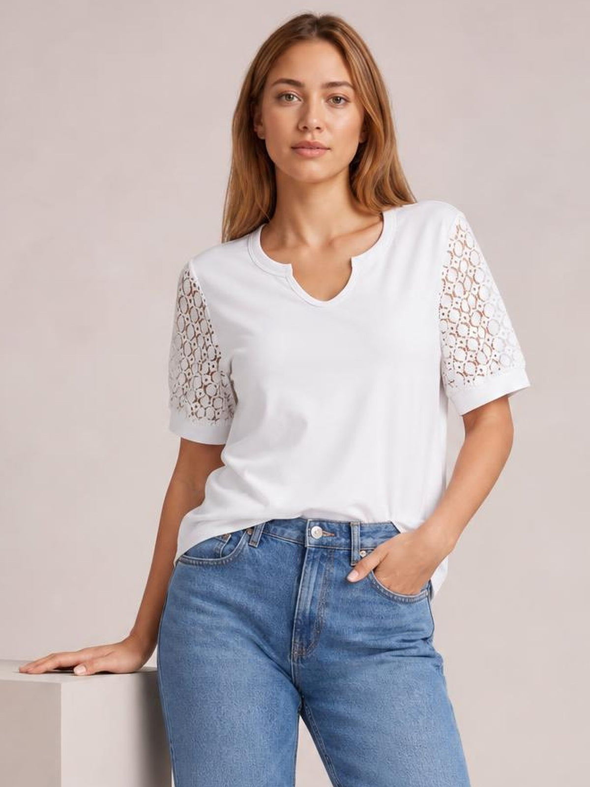 A model wears a white lace short sleeve T-shirt with semi-sheer openwork sleeves, a relaxed silhouette, and a notched neckline while standing with one hand in her pocket.
