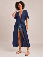 A model poses in a navy sheer beach cover-up maxi dress with a plunging V-neck, lace trim, short sleeves, tied waist, and flowing open-front silhouette.