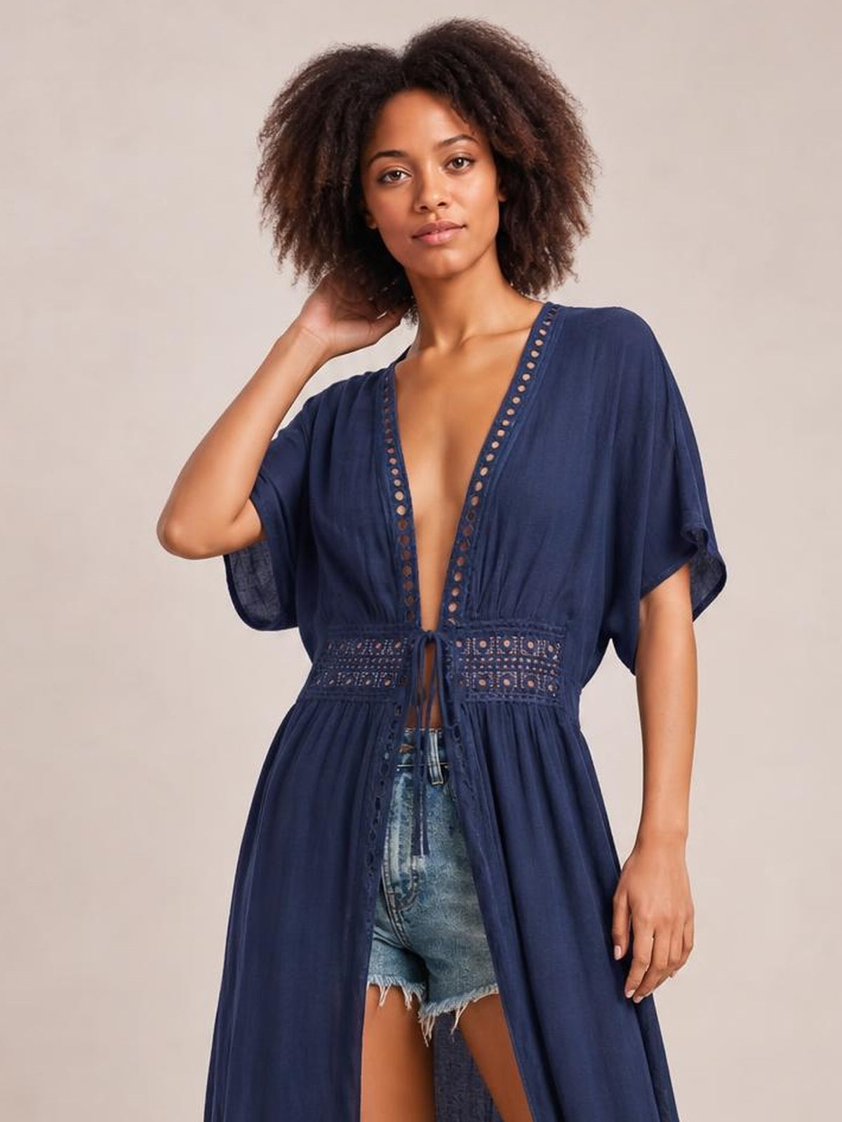 Model poses with one hand in her hair wearing a navy sheer plunging maxi beach cover-up with lace trim, short sleeves, a tied waist, and flowing silhouette.