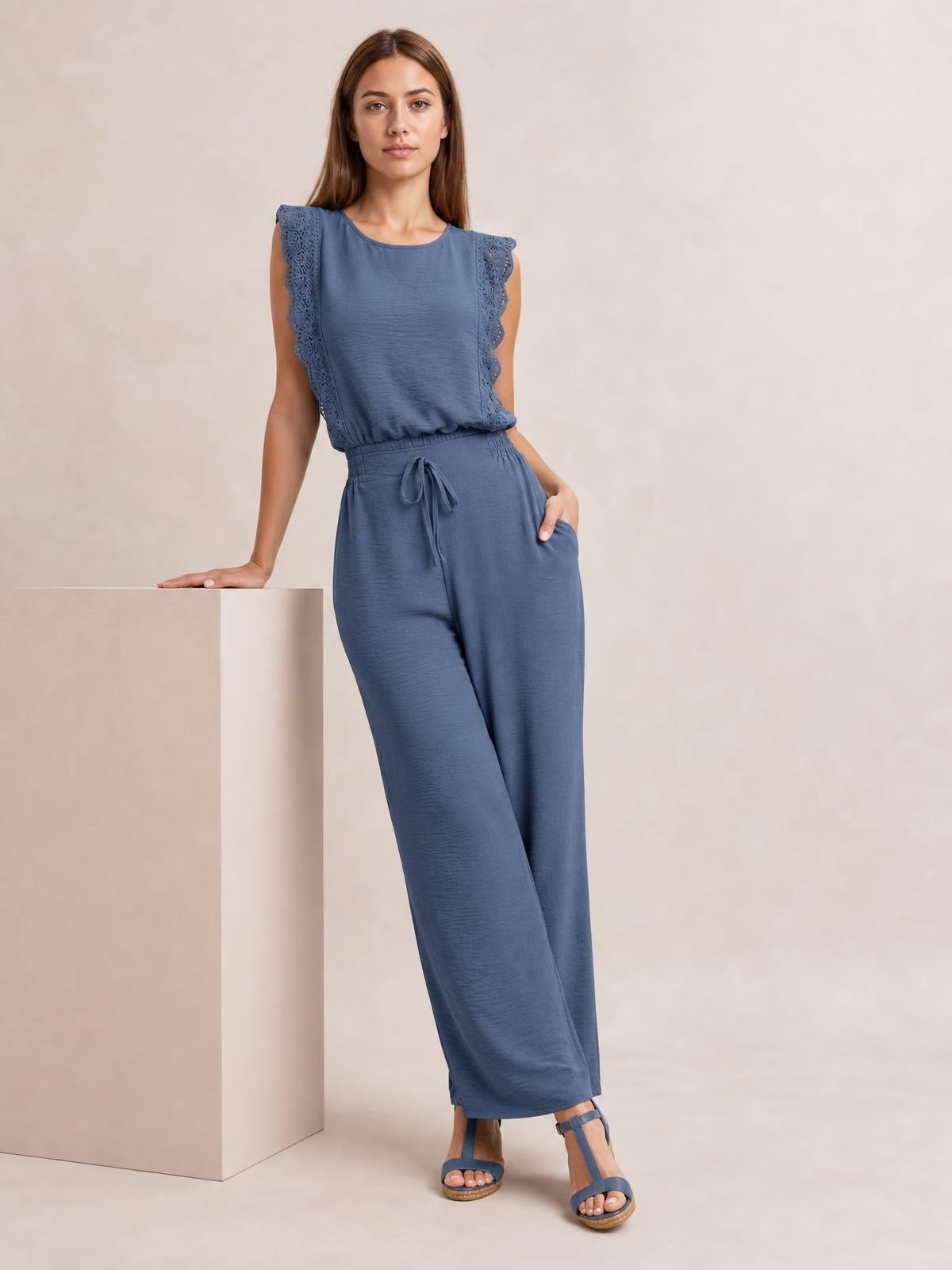 A model poses in a slate-blue lace-trim round-neck top and matching wide-leg pants set with tied waist, resting one hand on a pedestal.