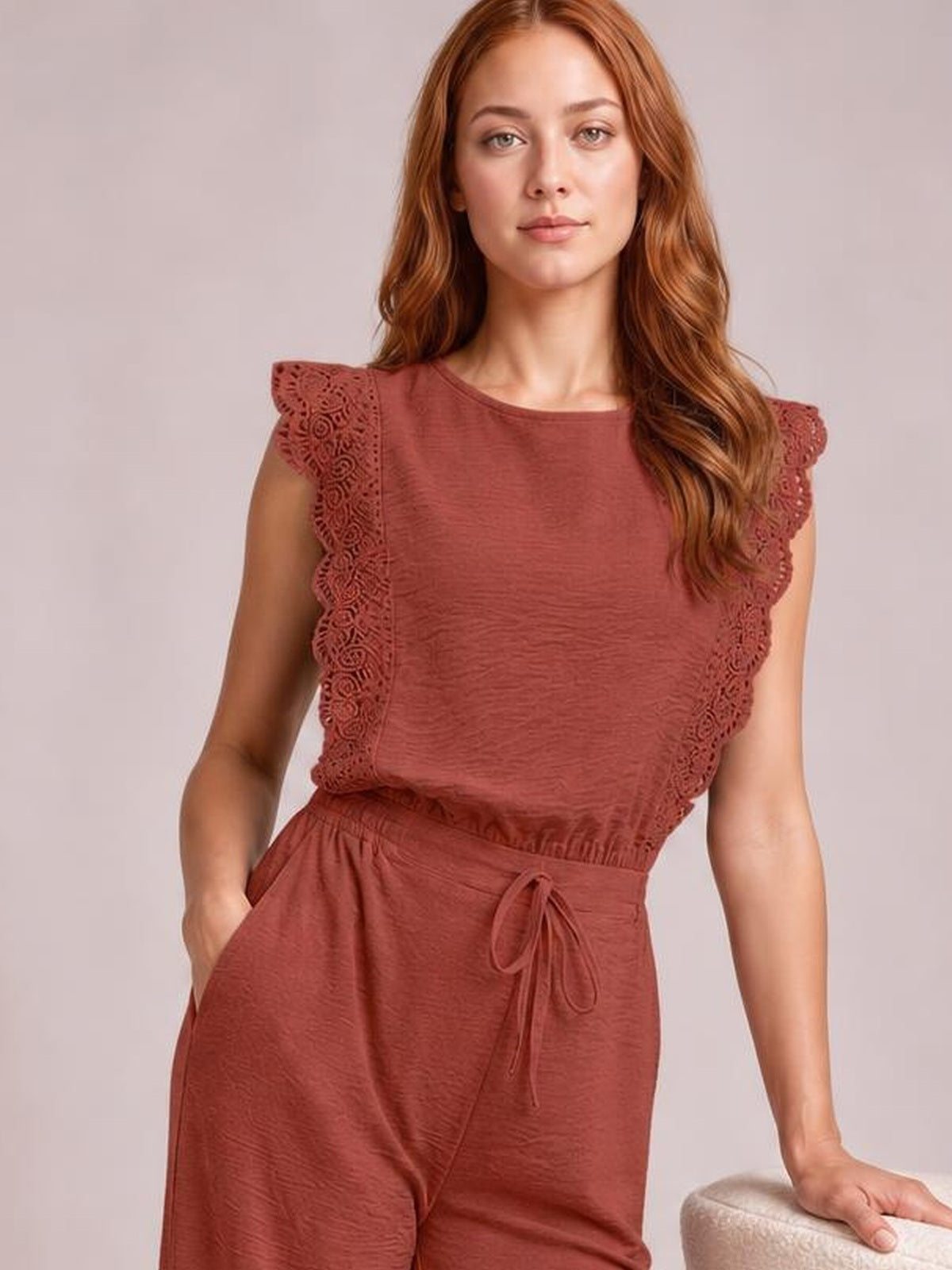 A model wears a brick-red round-neck top with scalloped lace-trim flutter sleeves and matching drawstring-waist pants, posing with one hand in a pocket.