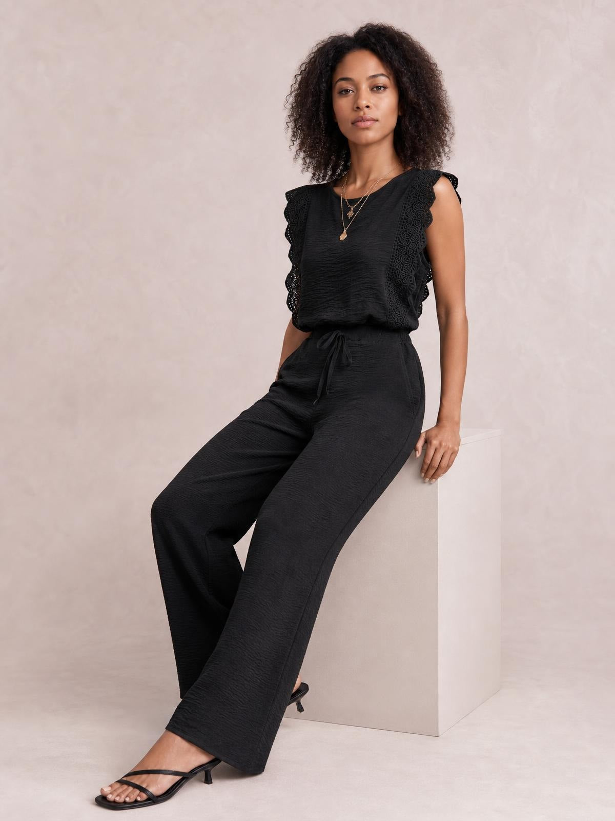A model poses seated in a black round-neck top and pants set with lace-trimmed shoulders, a tied waist, and wide-leg trousers.