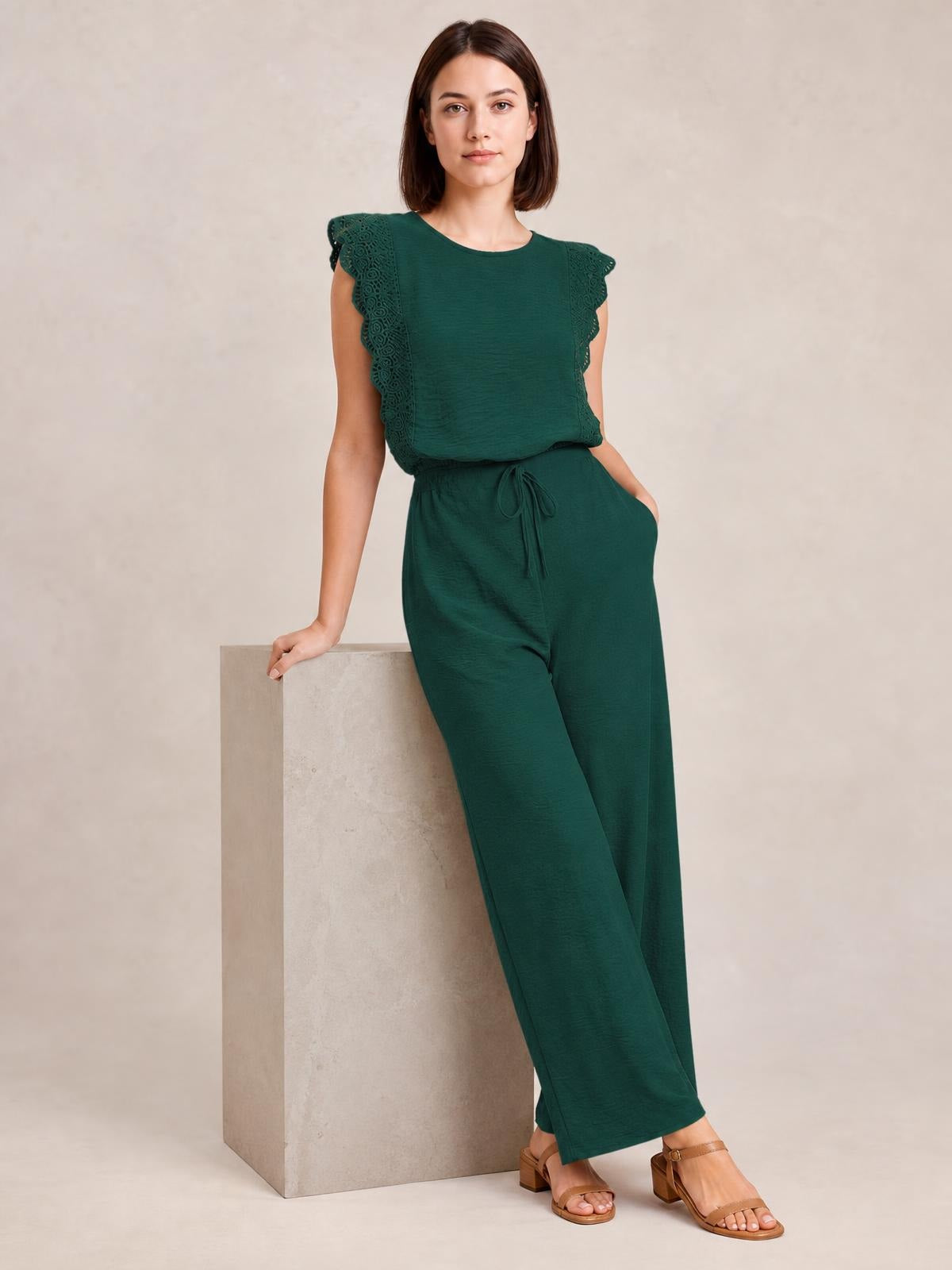 Model poses in a dark green round-neck top and pants set with scalloped lace-trim flutter sleeves, a tied waist, and relaxed wide-leg trousers.