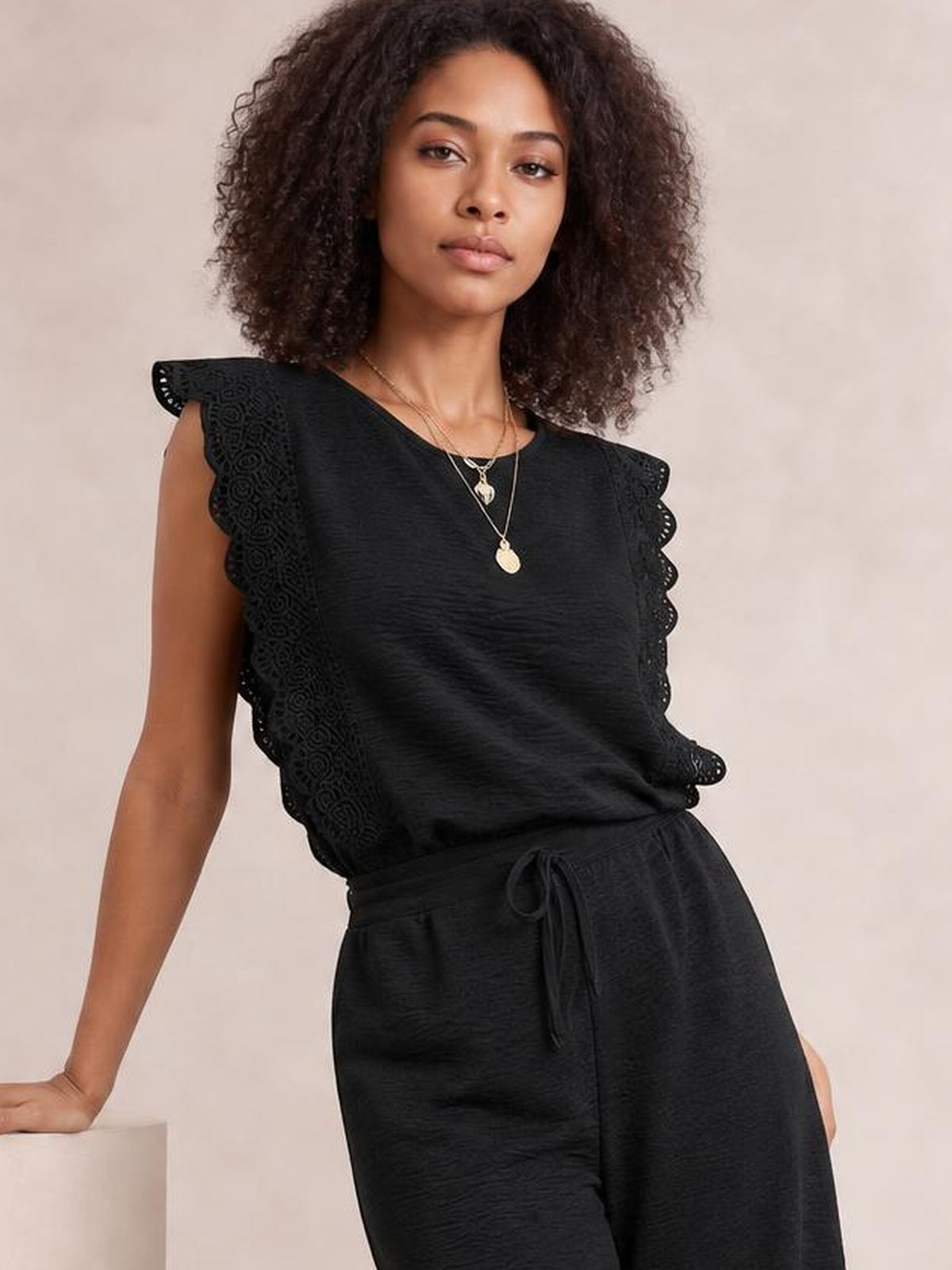Model poses in a black lace-trim round-neck top and matching pants set, featuring scalloped cap sleeves, a relaxed silhouette, and a tied waist.
