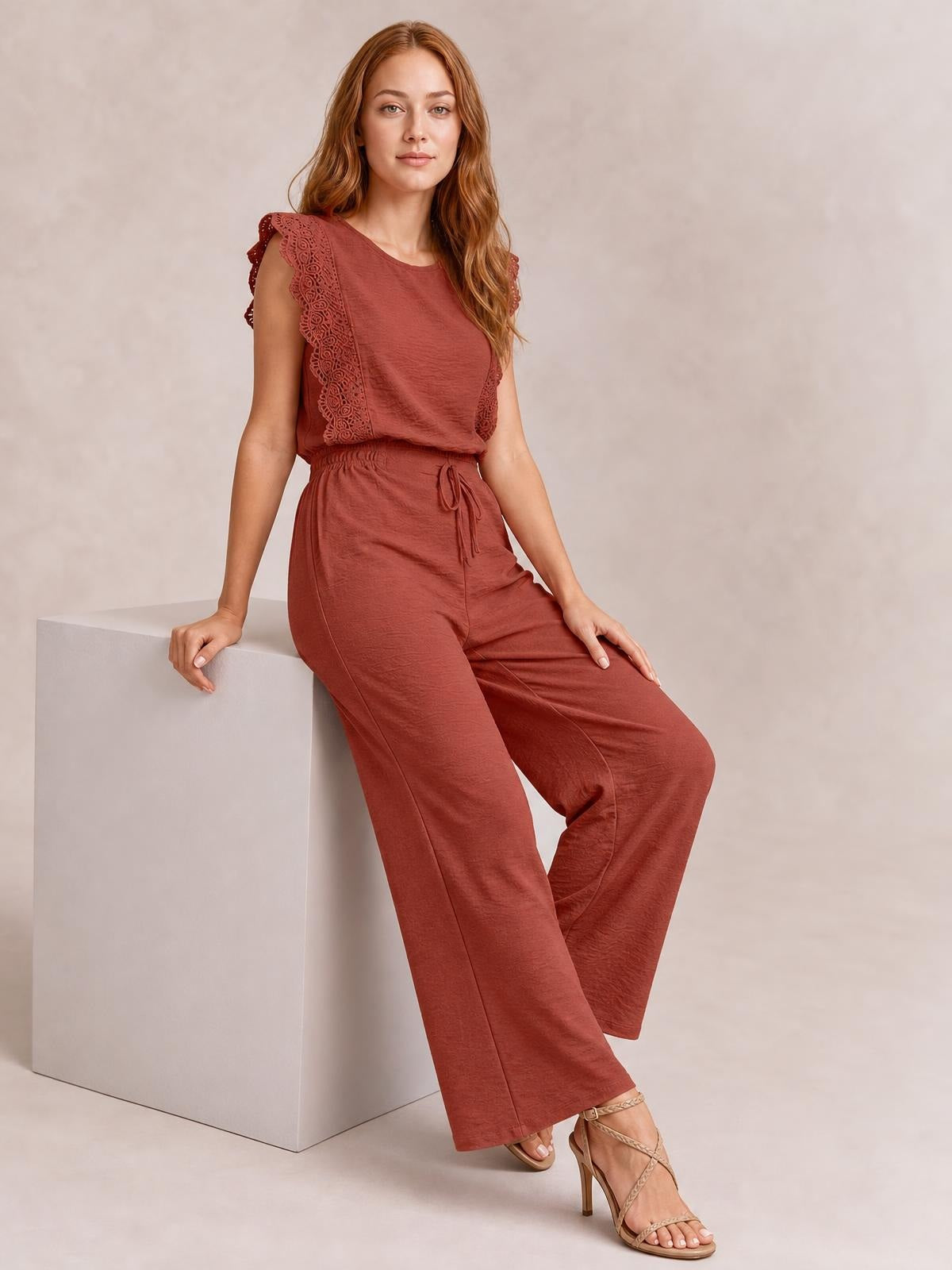 A seated model wears a rust-colored round-neck top with lace-trimmed flutter sleeves and matching wide-leg pants with a tied waist in a polished two-piece set.