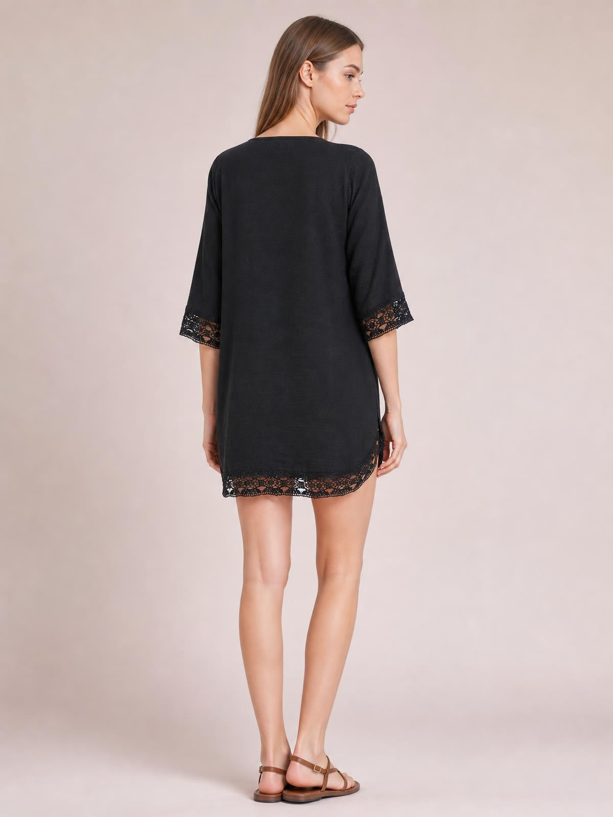 A model stands turned away in a black cover-up with a relaxed straight silhouette, three-quarter sleeves, and delicate lace trim at the cuffs and hem.