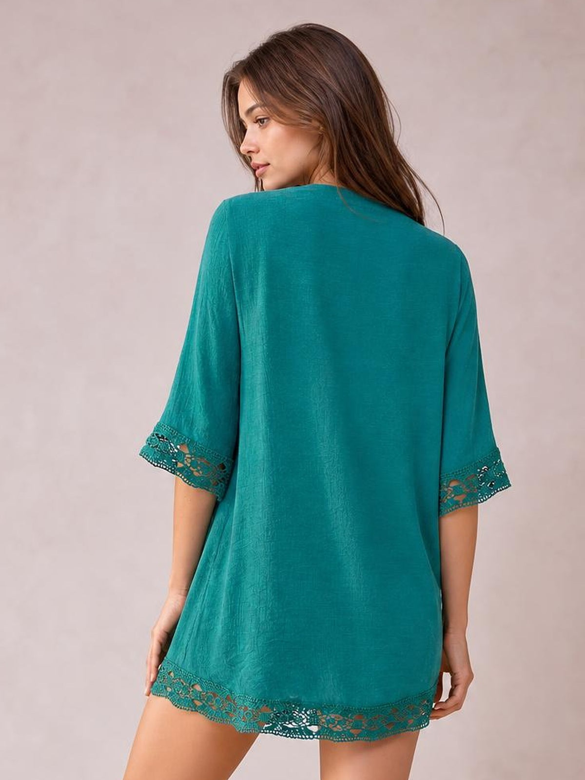 Model looks over her shoulder while wearing a teal cover-up with a loose silhouette, three-quarter sleeves, and delicate lace trim along the cuffs and hem.