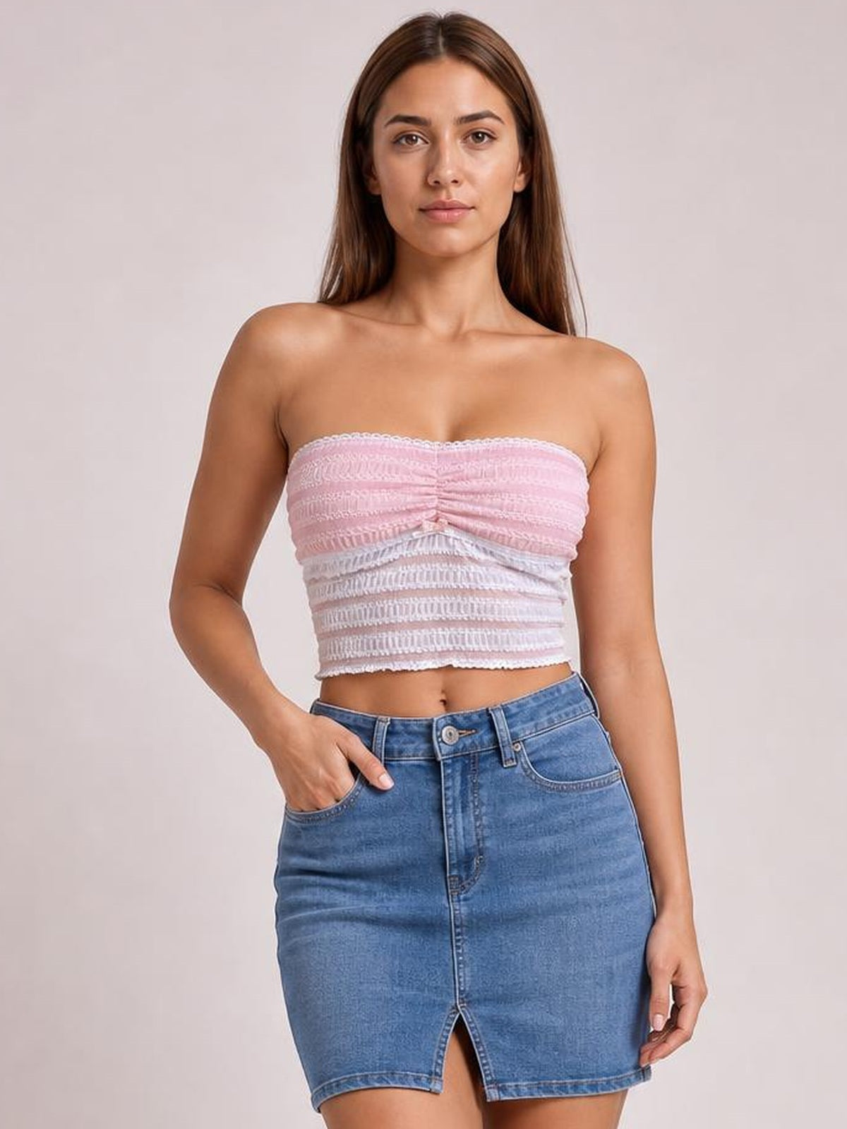 A model poses with one hand in her pocket wearing a pink lace tube top with a ruched center, strapless neckline, and fitted cropped silhouette.