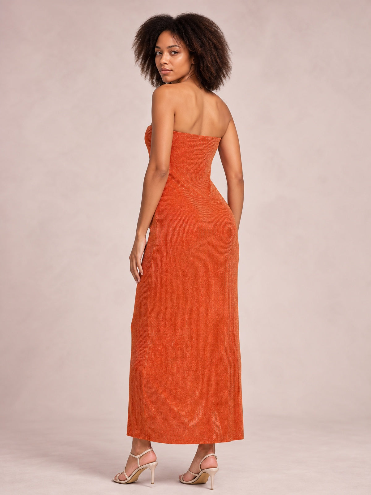 A model looks over her shoulder in an orange strapless maxi dress with a fitted column silhouette, textured fabric, and straight ankle-length hem.