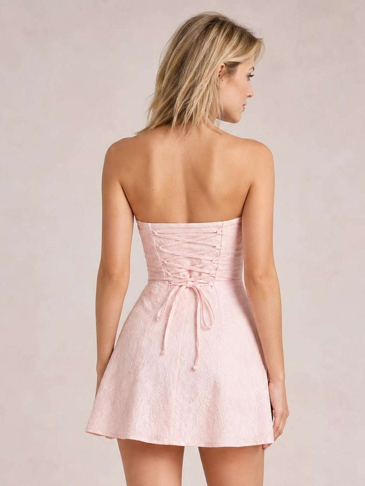 Model shown from the back in a pale pink lace-up jacquard tube mini dress with a fitted bodice, corset-style back, and flared skirt.