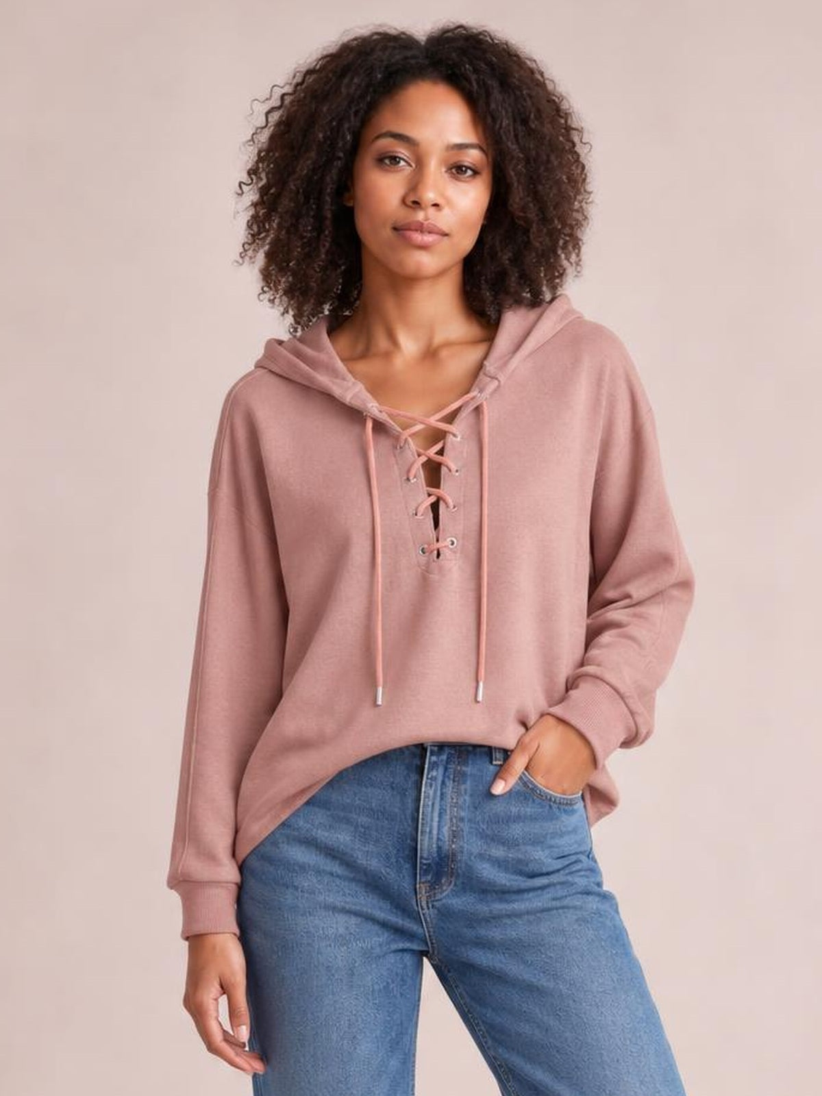 Model wearing a dusty rose lace-up long sleeve hoodie shirt with a relaxed silhouette, drawstrings, ribbed cuffs, and one hand in her jeans pocket.