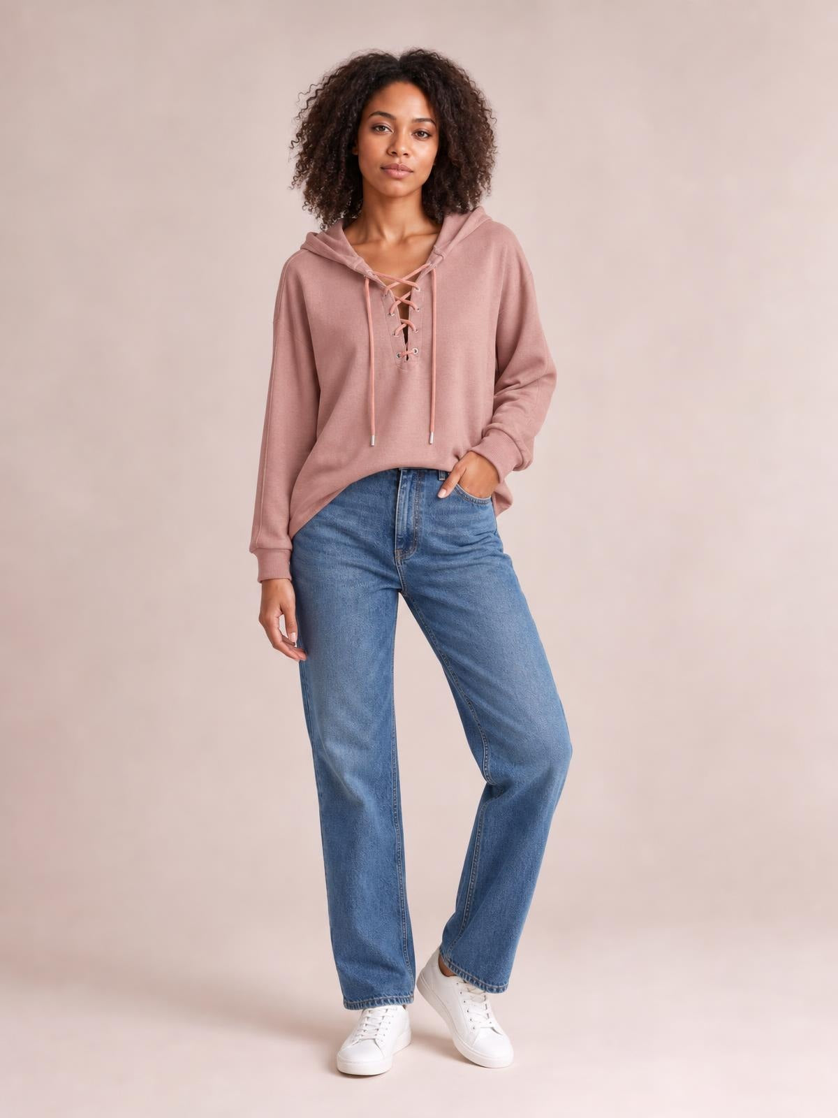 Model wears a dusty rose lace-up long sleeve hoodie with a relaxed silhouette, drawstring hood, ribbed cuffs, and a hand tucked into her jeans pocket.