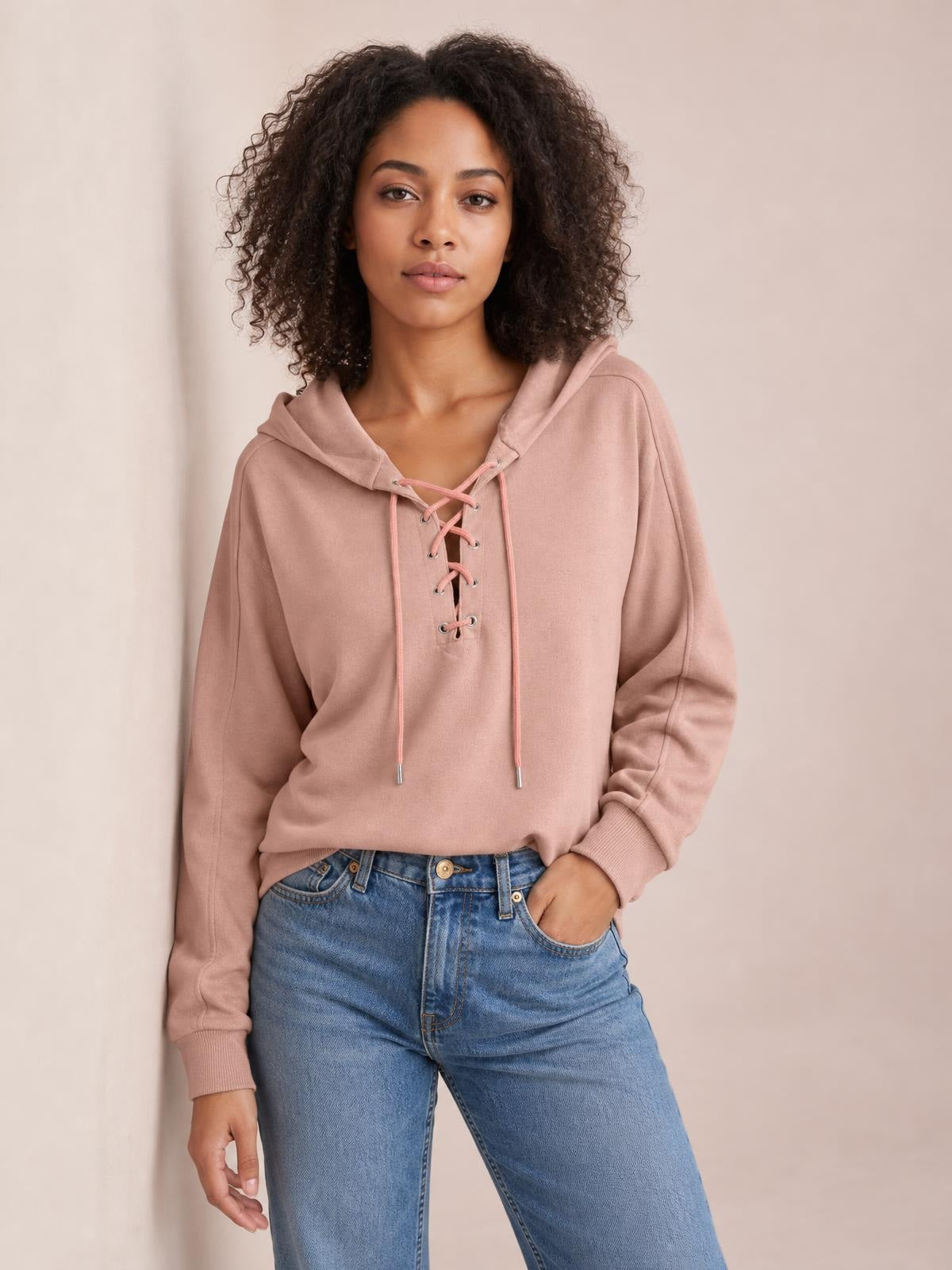 Model wears a dusty pink lace-up long sleeve hoodie with a drawstring hood, relaxed silhouette, ribbed cuffs, and one hand in her jeans pocket.