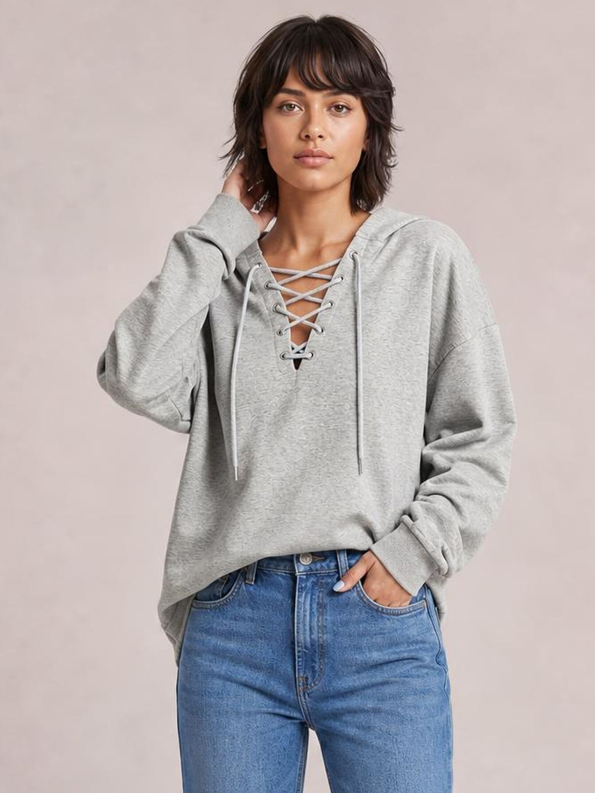 Model wears a light gray lace-up long sleeve hoodie with an oversized silhouette, drawstring hood, and crisscross neckline, posing with one hand in her jeans pocket.
