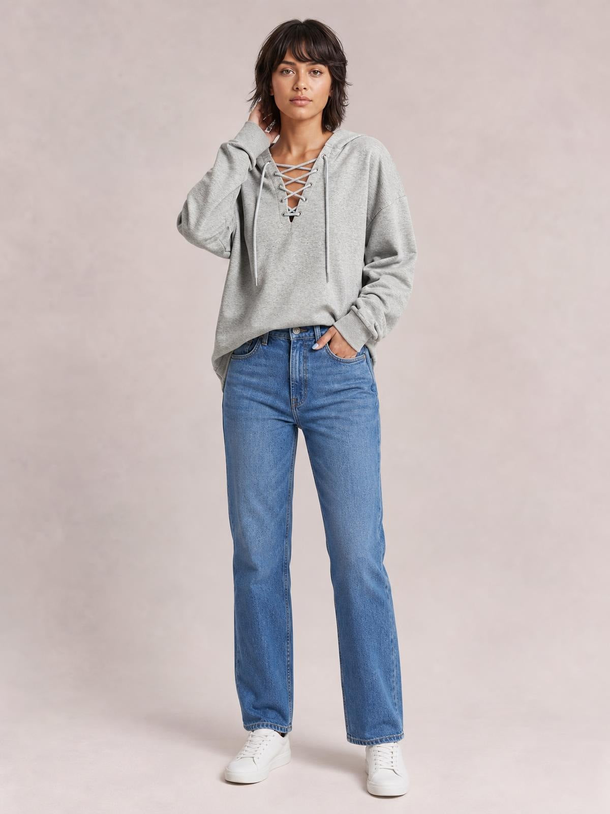 Model poses in a gray lace-up long sleeve hoodie with a relaxed silhouette, drawstring hood, and ribbed cuffs, one hand in her jeans pocket.
