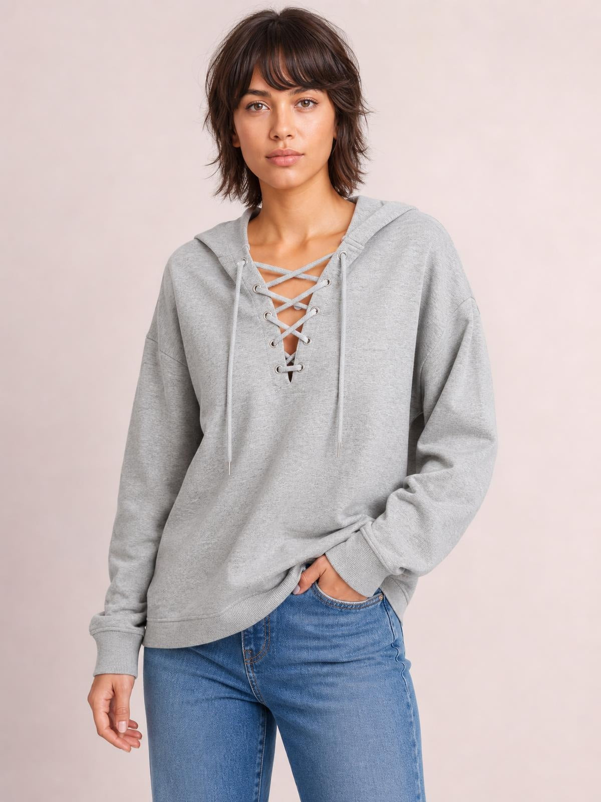 Model wearing a light gray long sleeve hoodie with a lace-up front, drawstring hood, relaxed silhouette, and ribbed cuffs, posed with one hand in her jeans pocket.