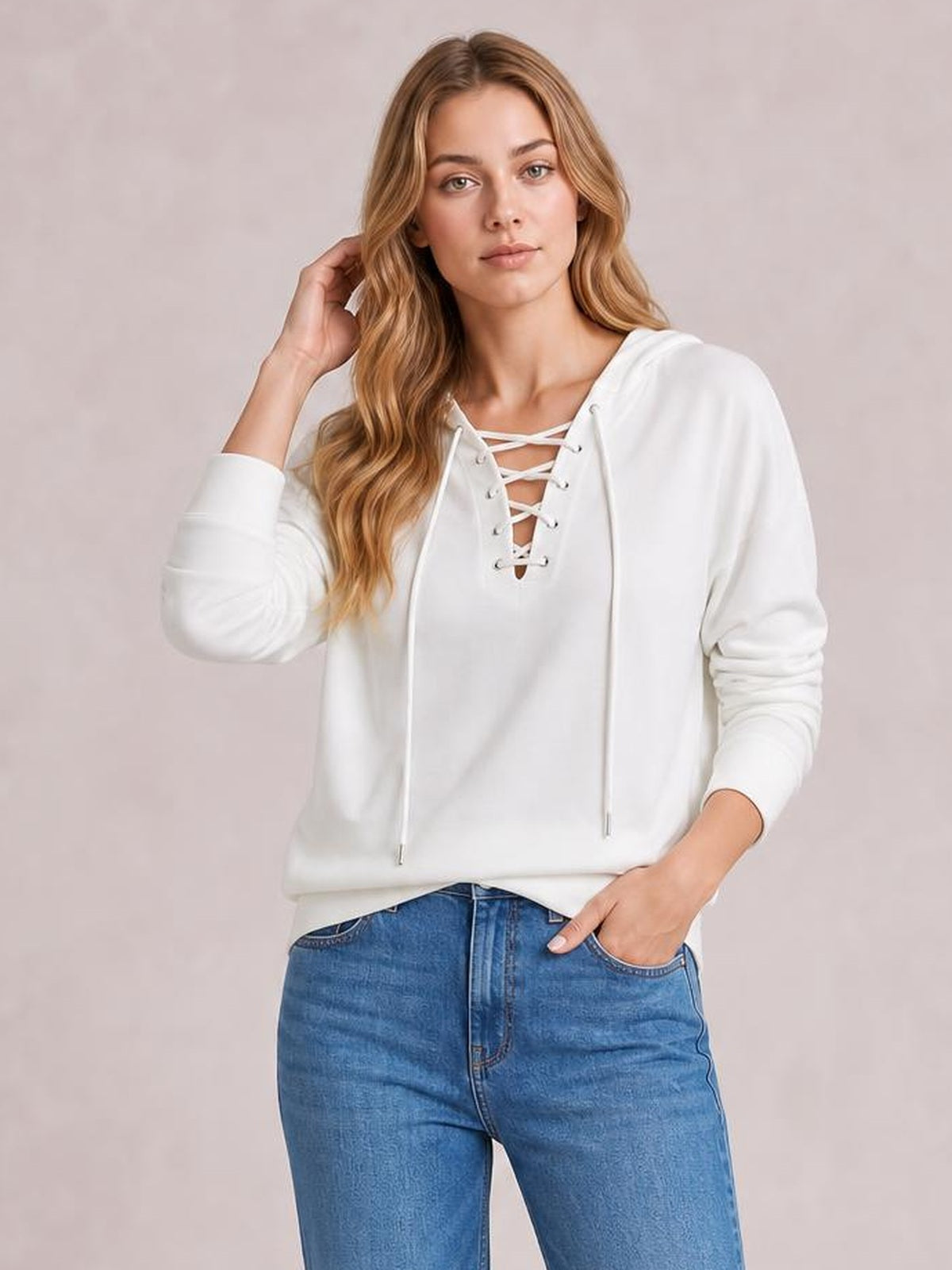 A model wears a white long-sleeve hoodie shirt with a lace-up front, relaxed silhouette, and drawstrings while touching her hair and resting one hand in her pocket.