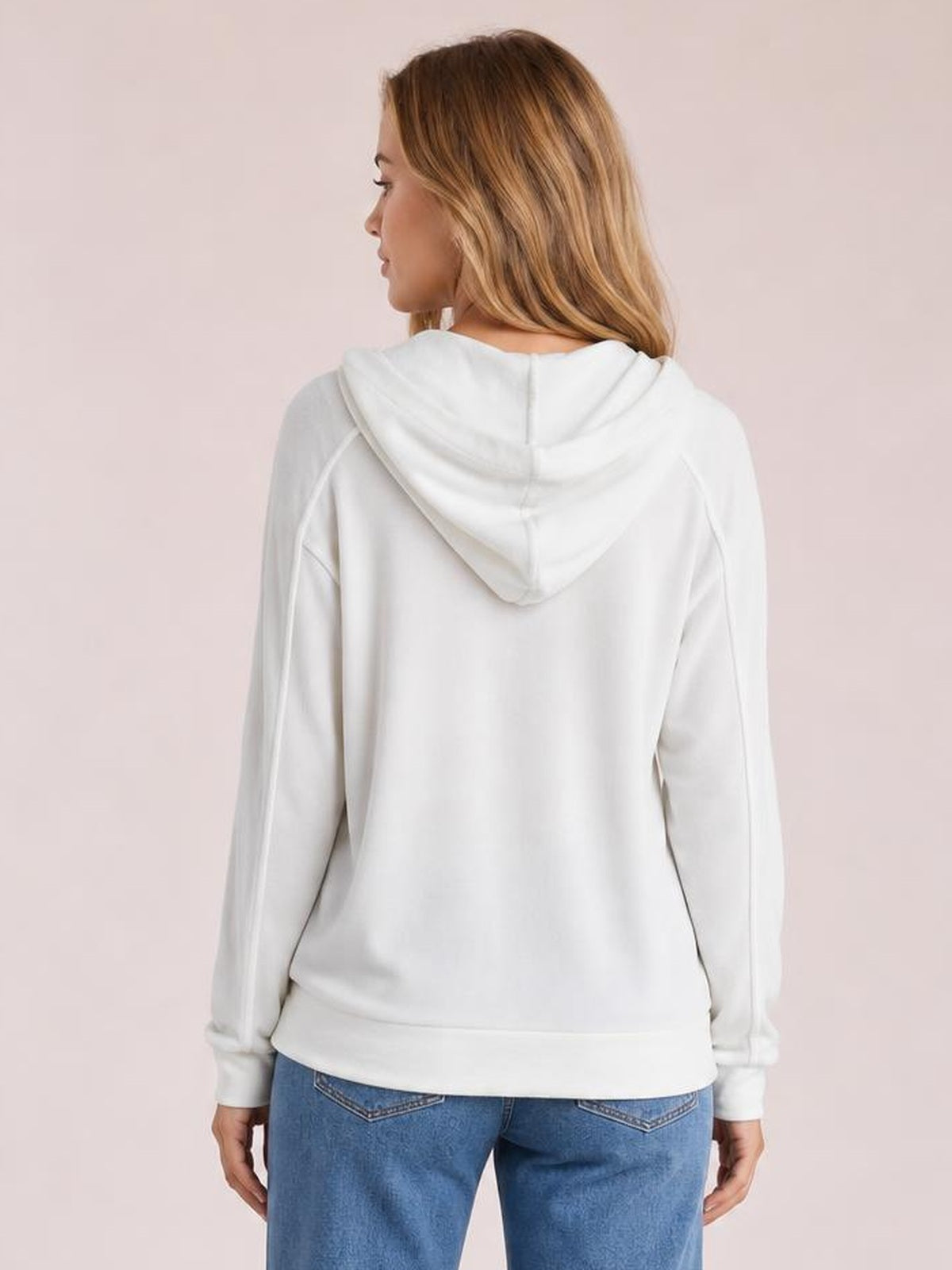 A model stands in a relaxed white long sleeve hoodie with a draped hood, raglan seams, ribbed cuffs, and a straight hem, shown from the back.