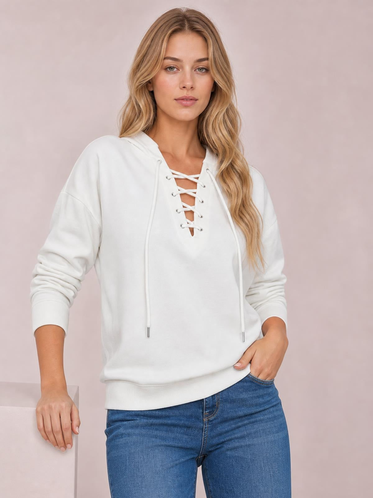 Model wears a white lace-up long sleeve hoodie with a relaxed silhouette, hood, ribbed cuffs and hem, posing with one hand in her jeans pocket.
