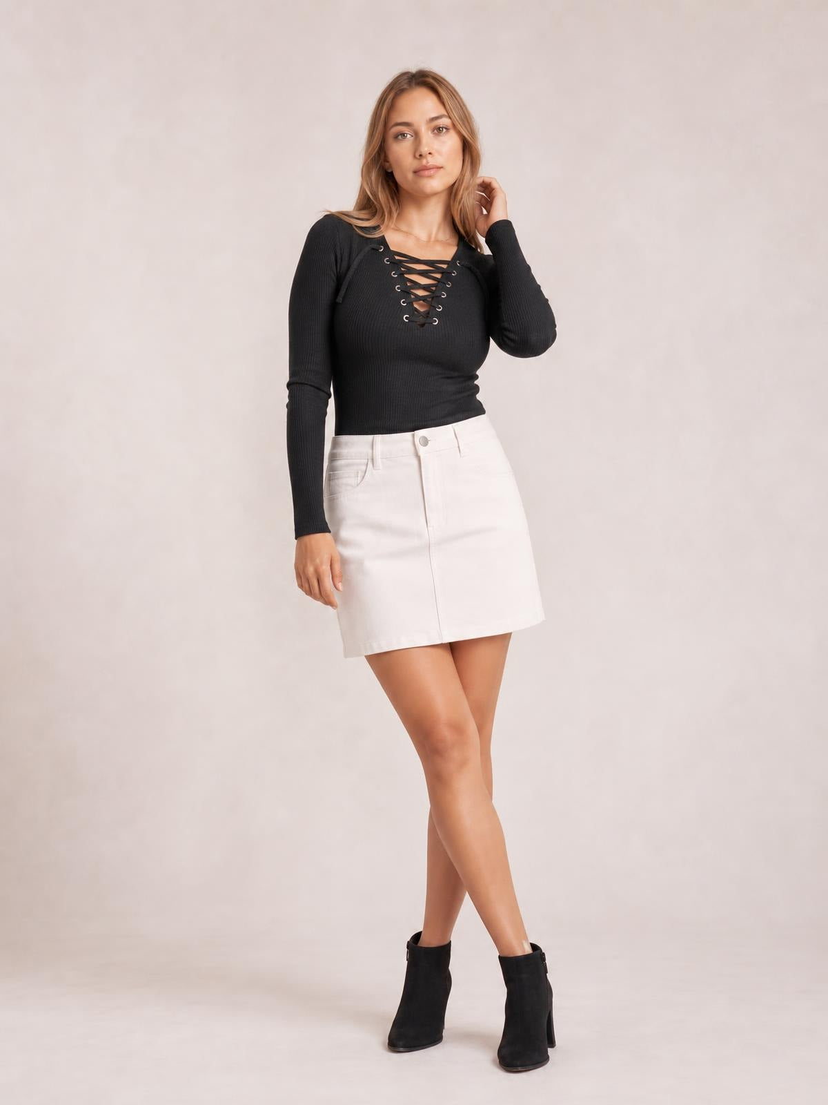 Model poses in a fitted black ribbed lace-up long sleeve T-shirt with crisscross chest detail, paired with a white skirt.
