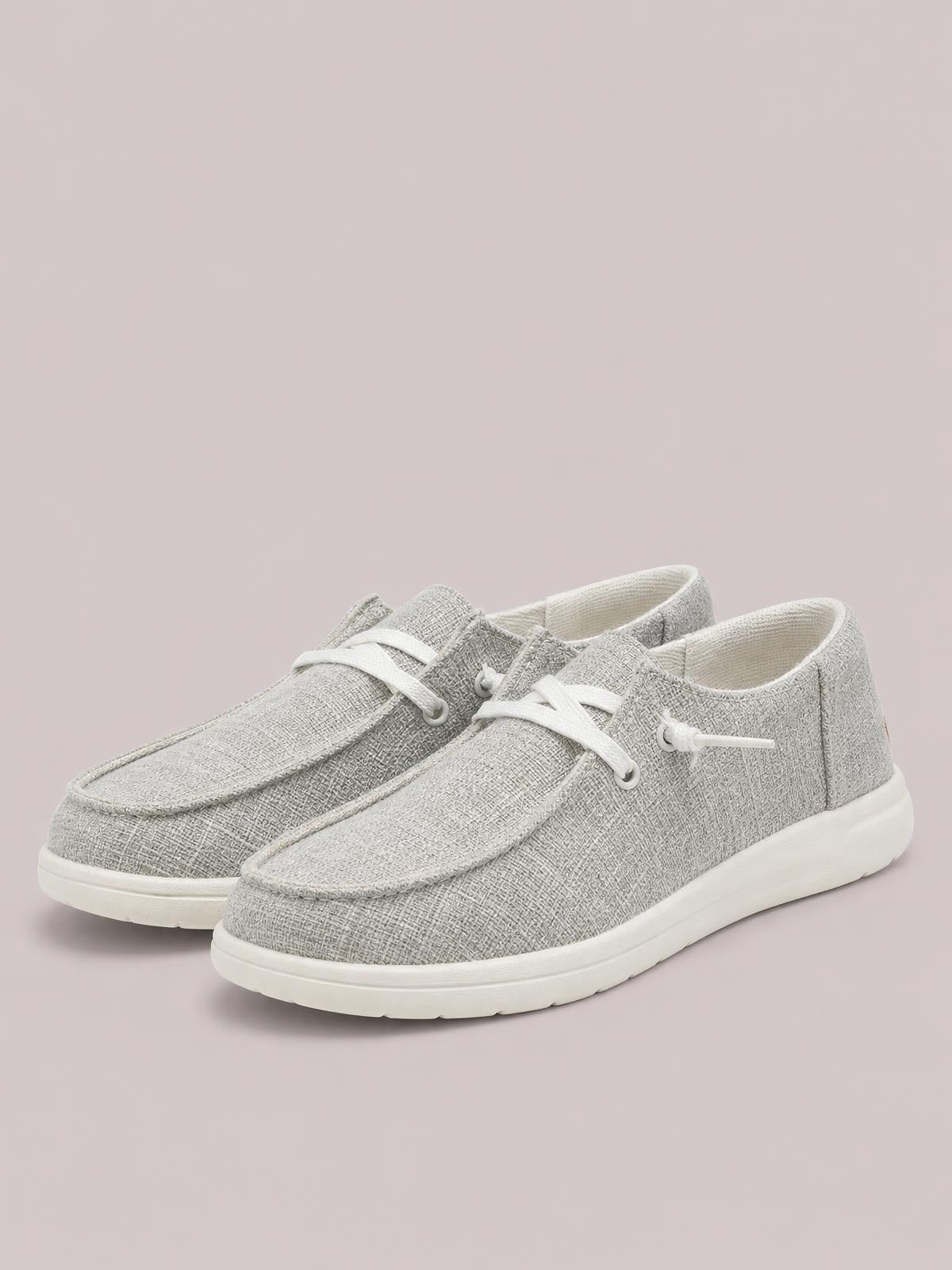 Light gray canvas lace-up slip-ons feature rounded toes, white laces, stitched paneling, and thick flat rubber soles.