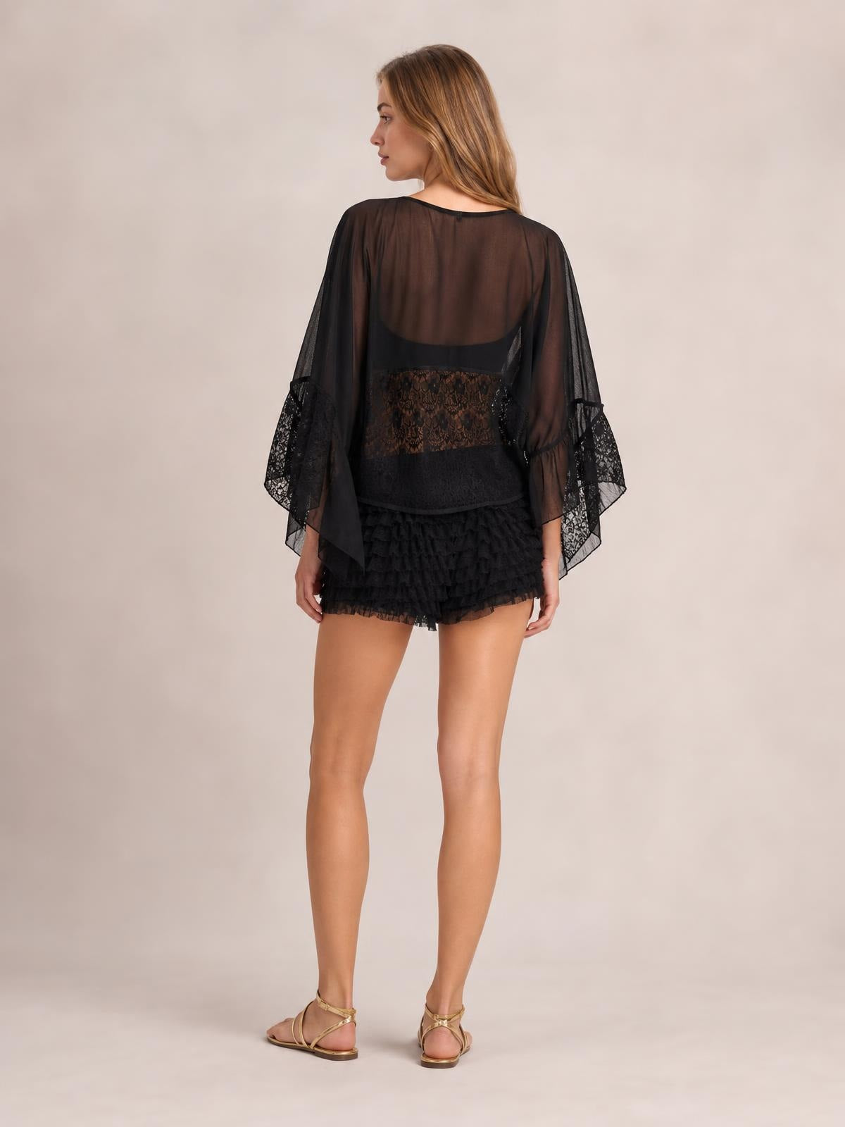 A model stands facing away in a black sheer lace long-sleeve cover-up with flowing sleeves, ruffled lace cuffs, and a relaxed silhouette.