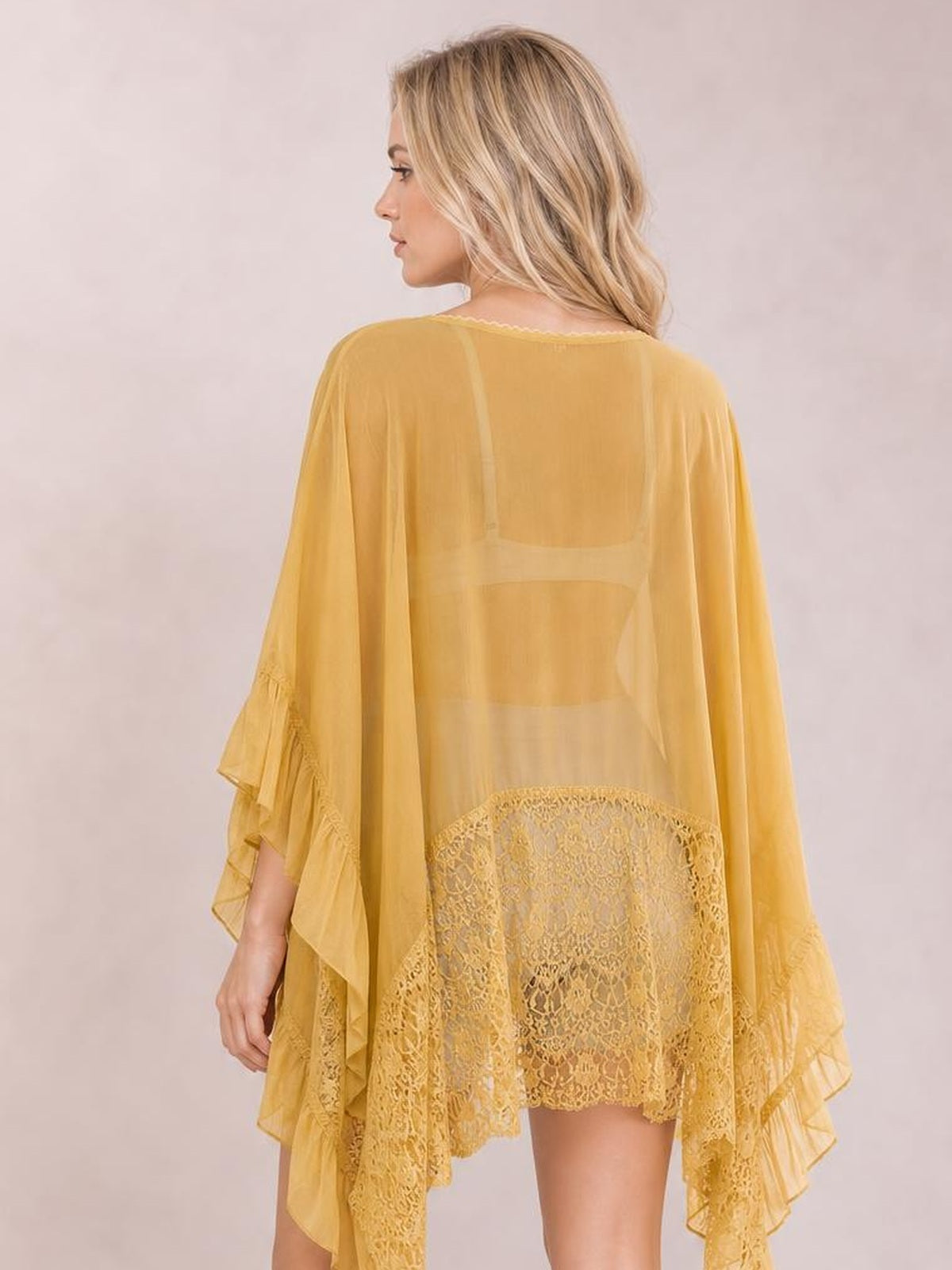 A model poses from behind in a mustard yellow lace cover-up with sheer long sleeves, a flowing silhouette, ruffled edges, and a delicate lace hem.