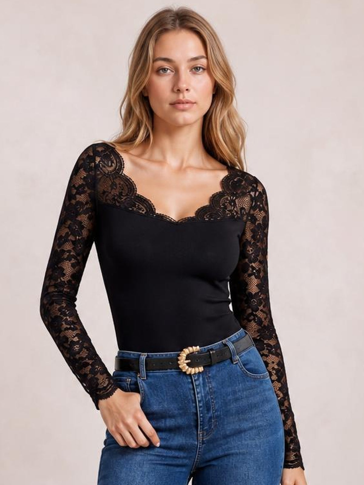 A model poses in a fitted black lace top with semi-sheer long sleeves, a scalloped V-neckline, and one hand resting in her jeans pocket.