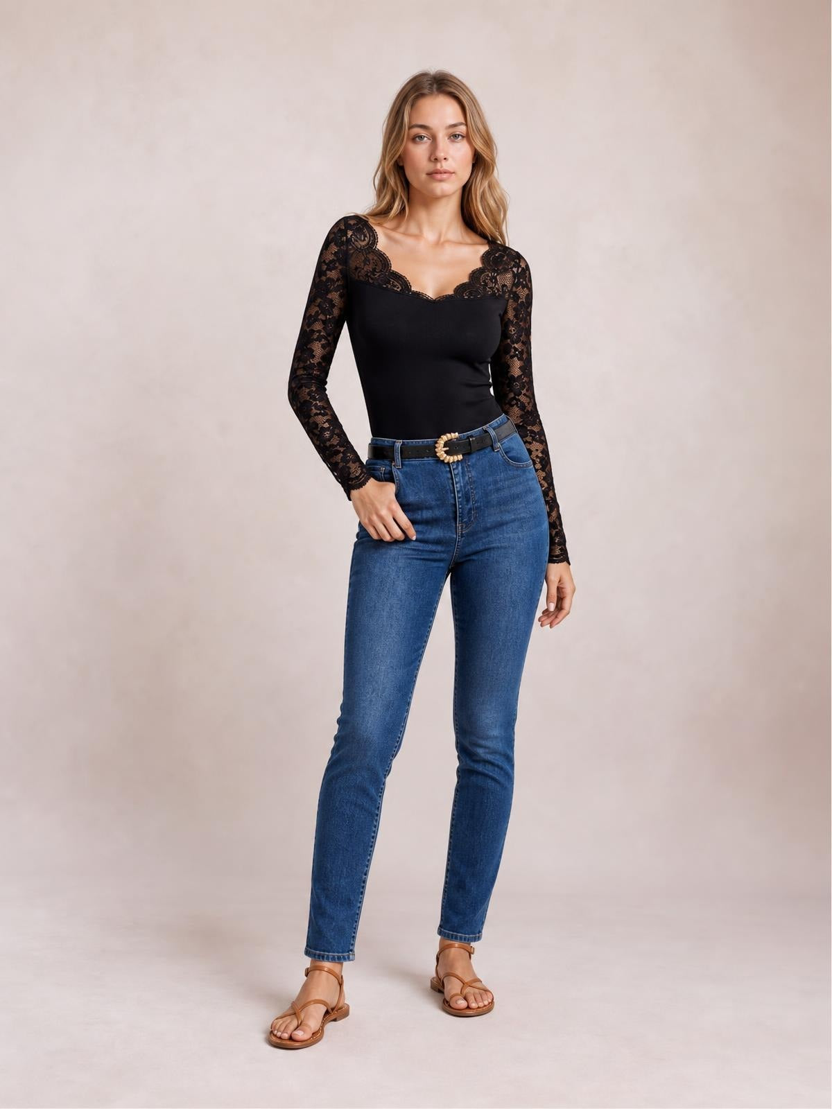Model wearing a black semi-sheer lace long-sleeve top with scalloped wavy hem and V-neckline, styled in a fitted silhouette with one hand on her hip.