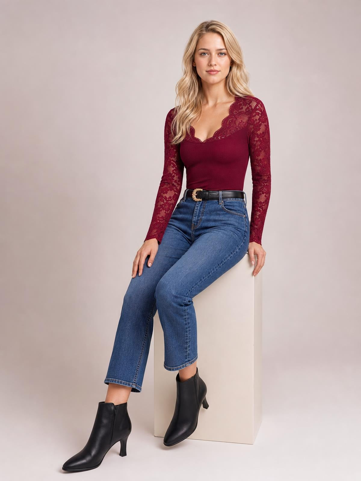 A seated model wears a burgundy lace V-neck long sleeve top with semi-sheer sleeves, a fitted silhouette, and a softly scalloped neckline.