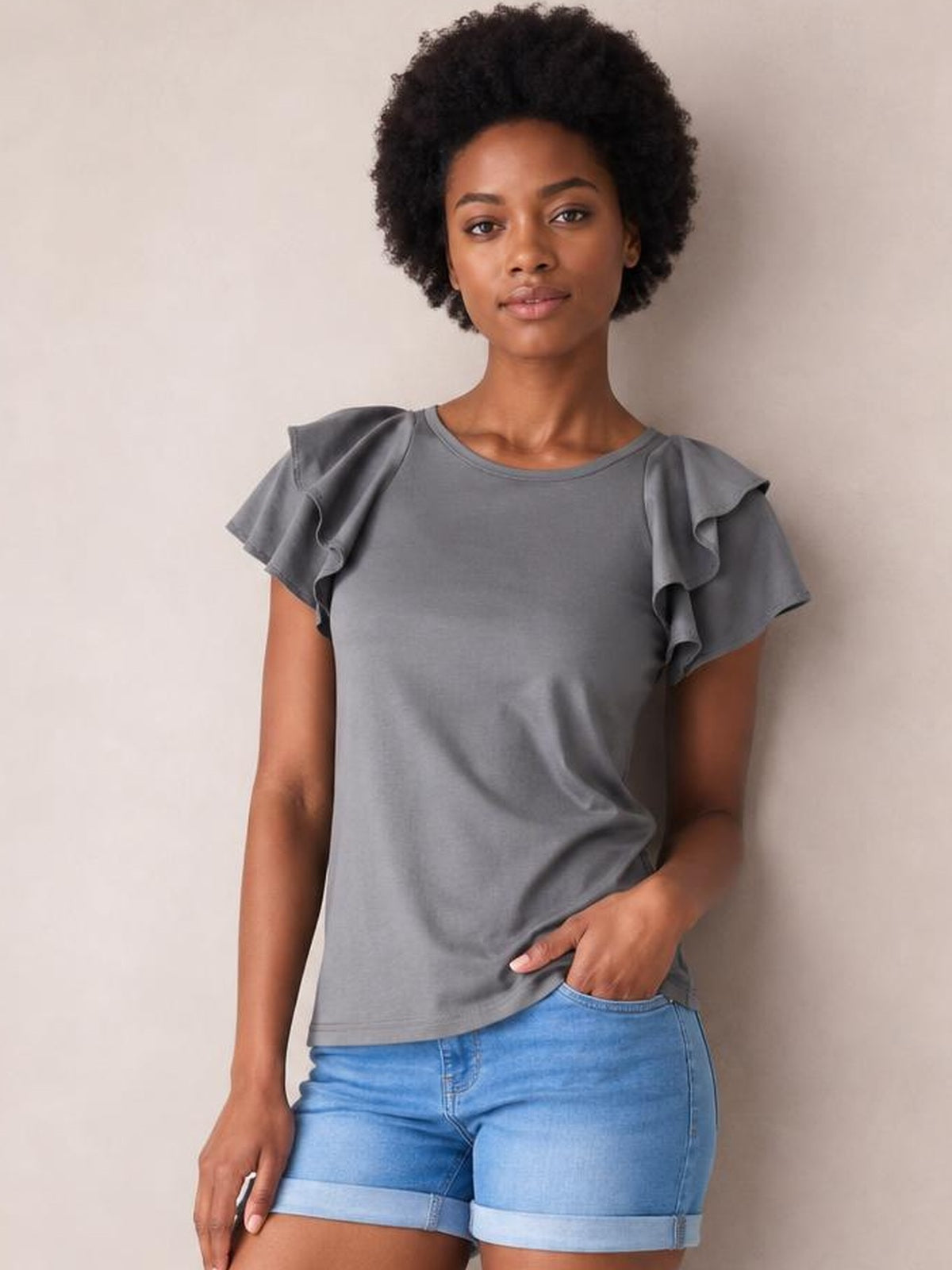 Model wearing a solid gray layered flutter sleeve round neck top with a regular silhouette, posing with one hand in her denim shorts pocket.