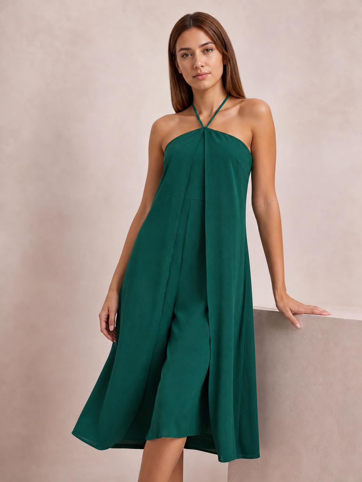 Model standing in a deep green layered halter neck wide-leg jumpsuit with flowing panels and a relaxed, elegant silhouette.