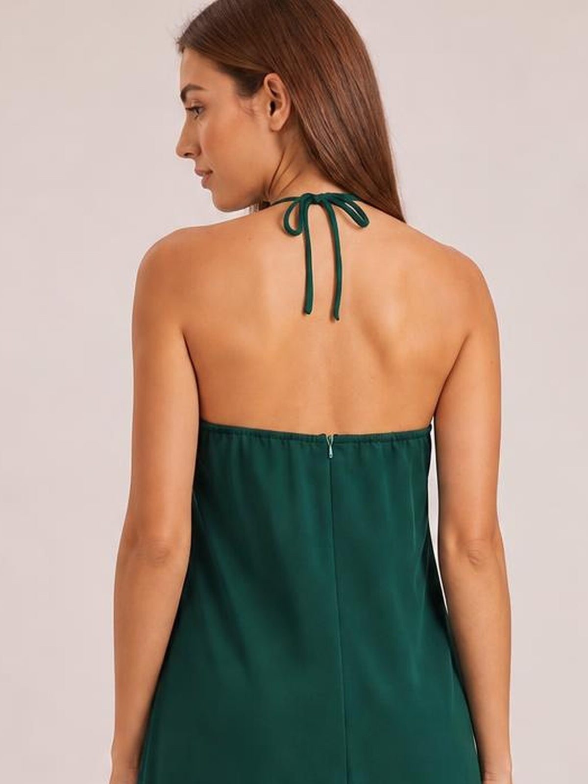 A model viewed from behind wears a dark green halter-neck jumpsuit with a tied neck, exposed back, center-back zipper, and wide-leg silhouette.