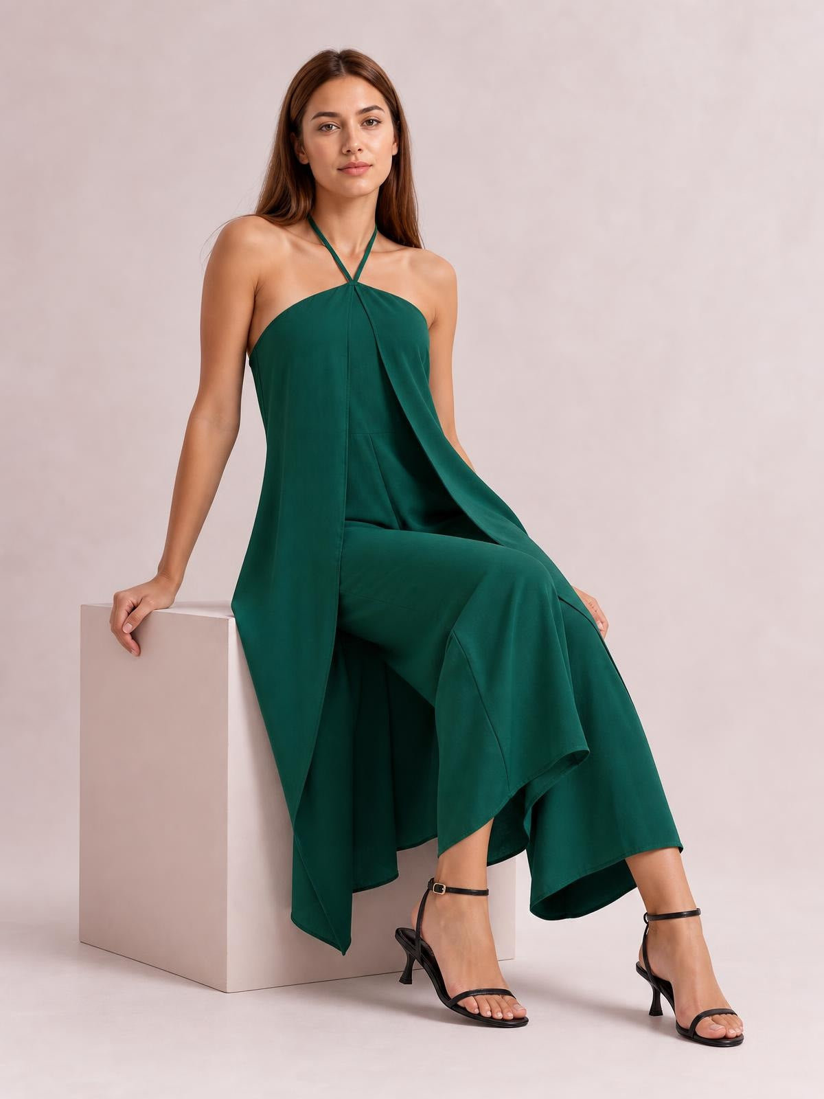 A model sits in a deep green layered halter neck jumpsuit with a draped overlay bodice and flowing wide-leg silhouette.
