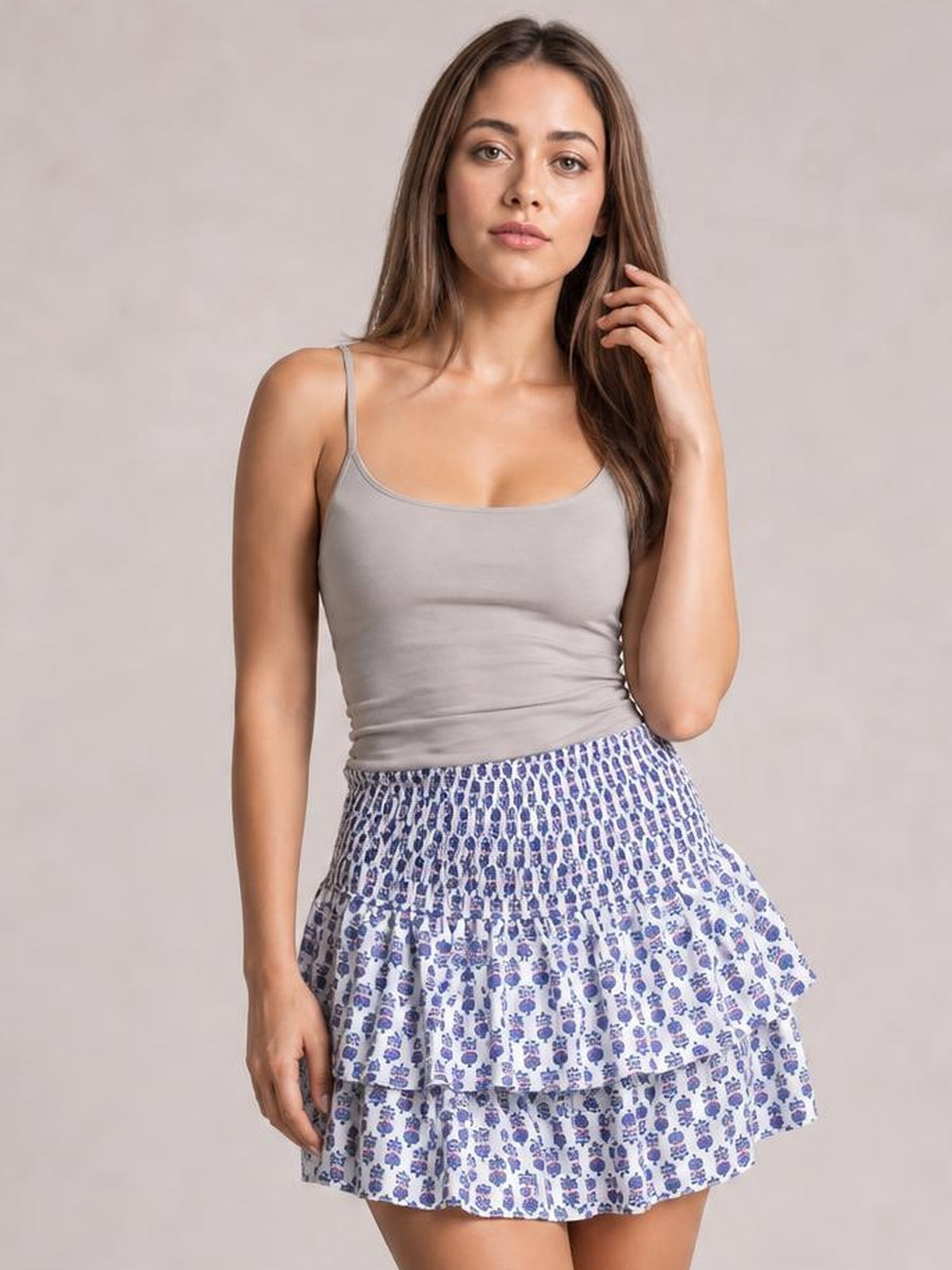 Model wearing a blue-and-white printed layered mini skirt with a smocked waistband, ruched tiers, and ruffled flared silhouette, posing with one hand in her hair.