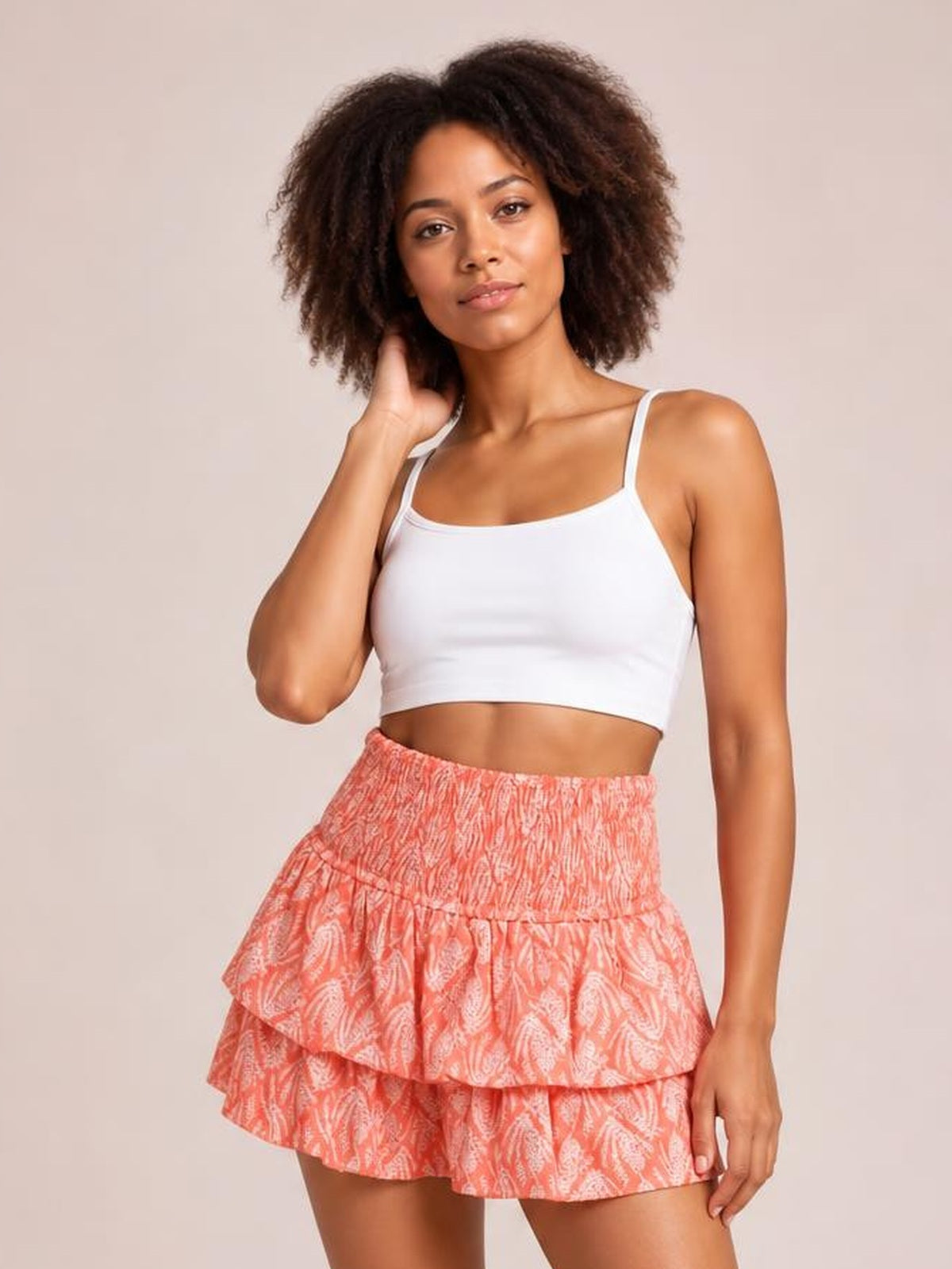 Model poses with one hand in her hair wearing a coral patterned layered mini skirt with a smocked waist and tiered ruched ruffles.