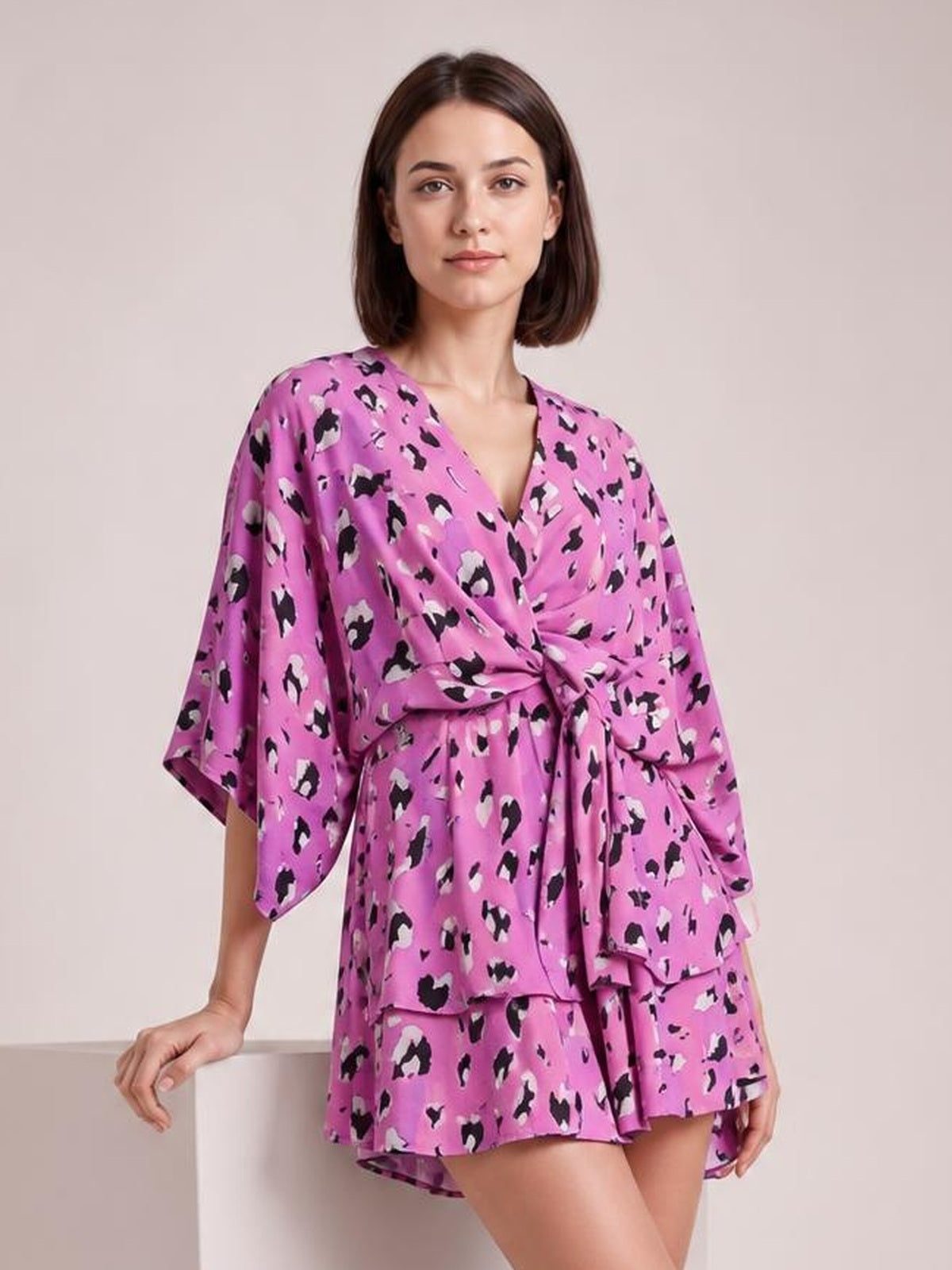 A model poses in a pink printed surplice mini dress with a V-neck, wide three-quarter sleeves, ruched tie waist, and layered tiered skirt.