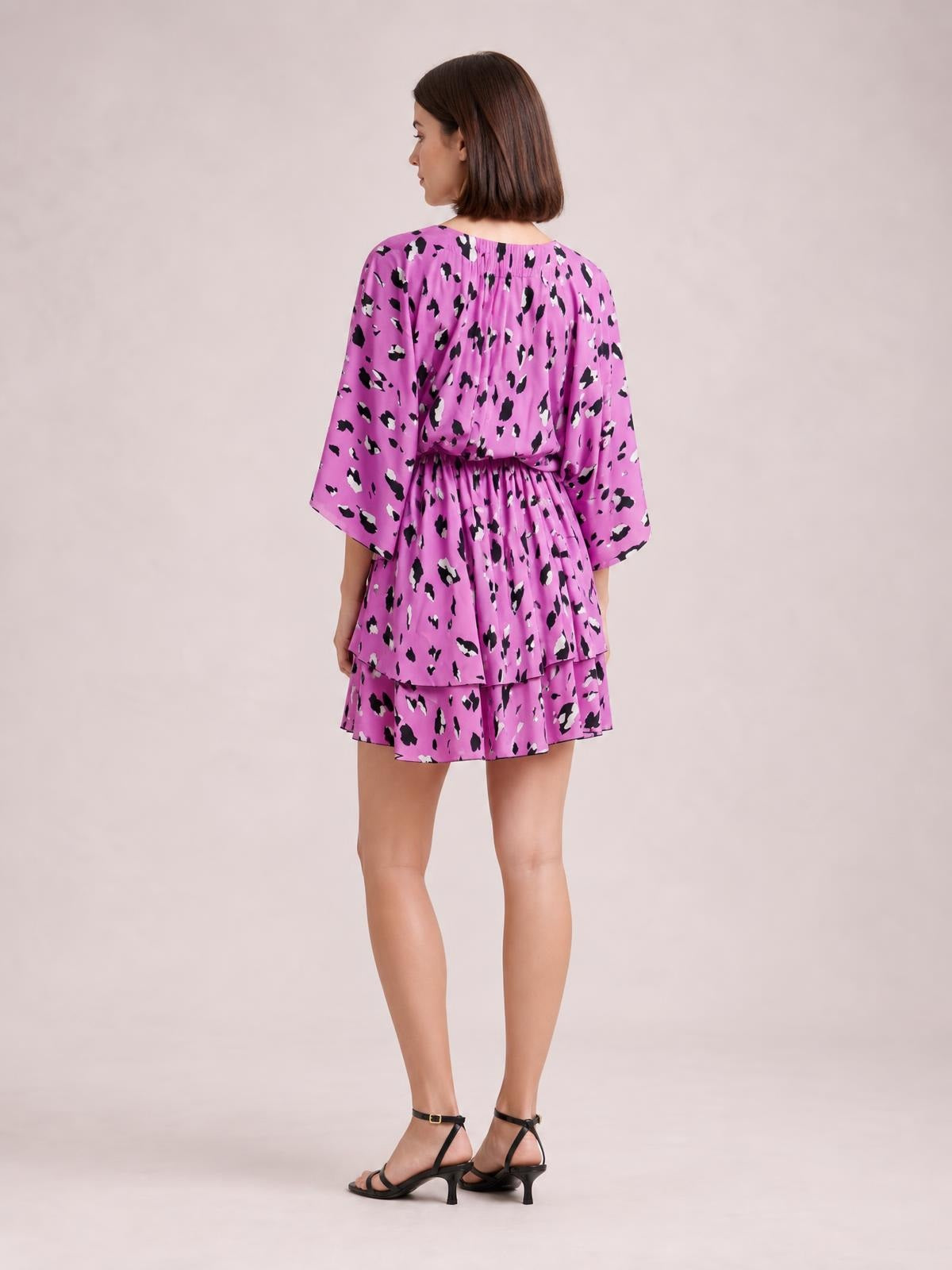 Model shown from the back in a bright pink printed mini dress with long sleeves, a ruched waist, and layered tiered ruffle hem.
