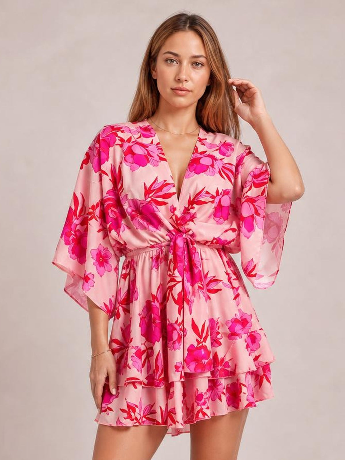 Model poses in a pink floral printed surplice mini dress with wide sleeves, a tied waist, ruched detail, and tiered ruffle skirt.
