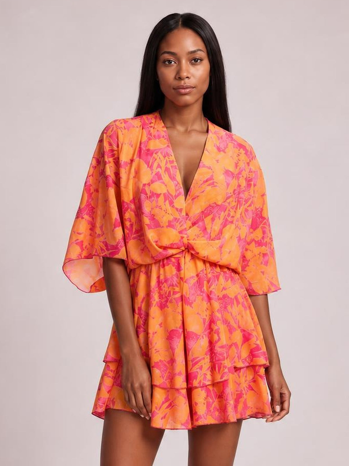 Model poses in a vibrant orange and pink printed surplice mini dress with a belted waist, wide sleeves, ruched front, and tiered flared skirt.