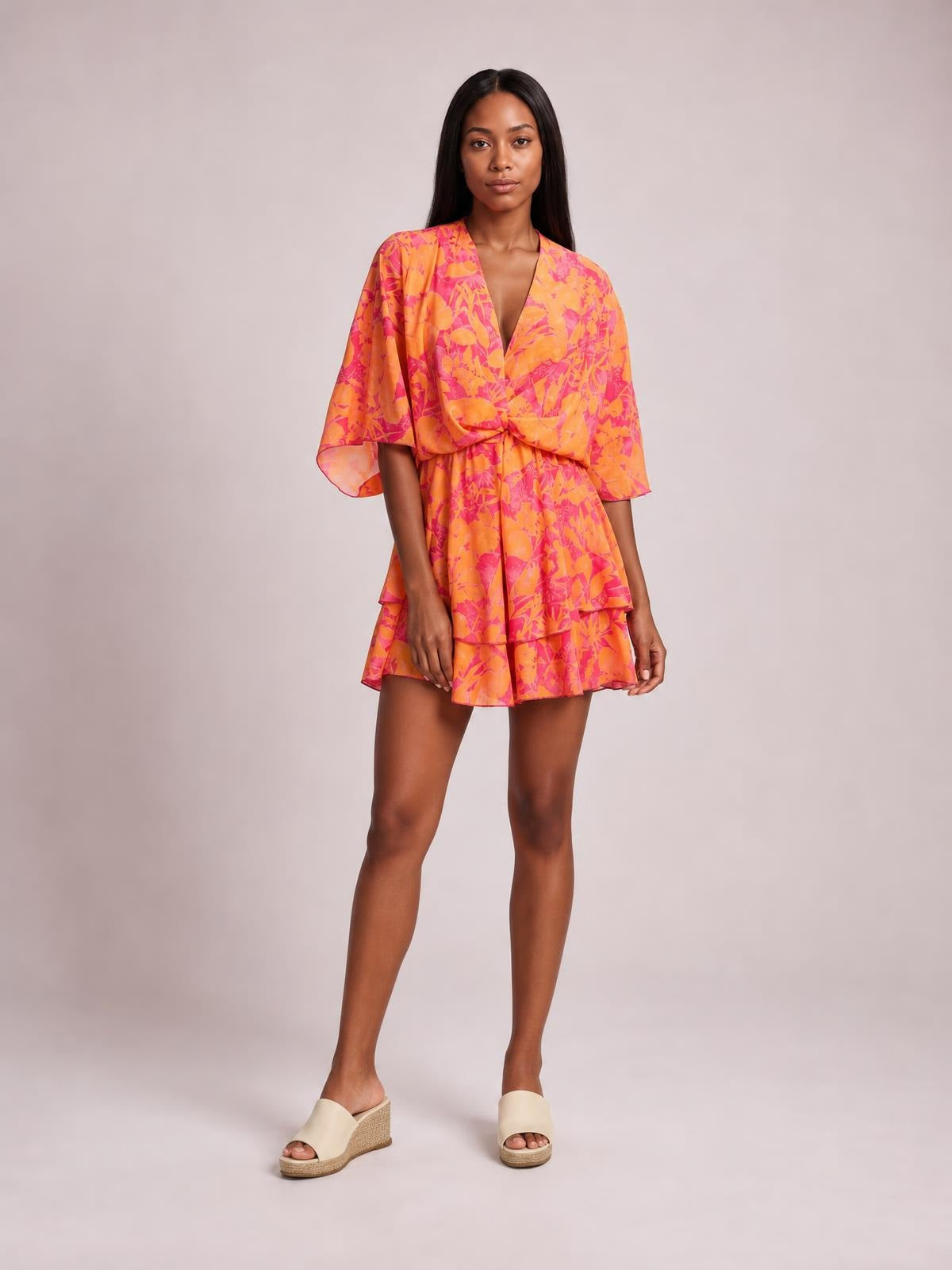 Model stands in a bright orange and pink printed surplice mini dress with loose sleeves, a ruched tie waist, and a tiered ruffled skirt.